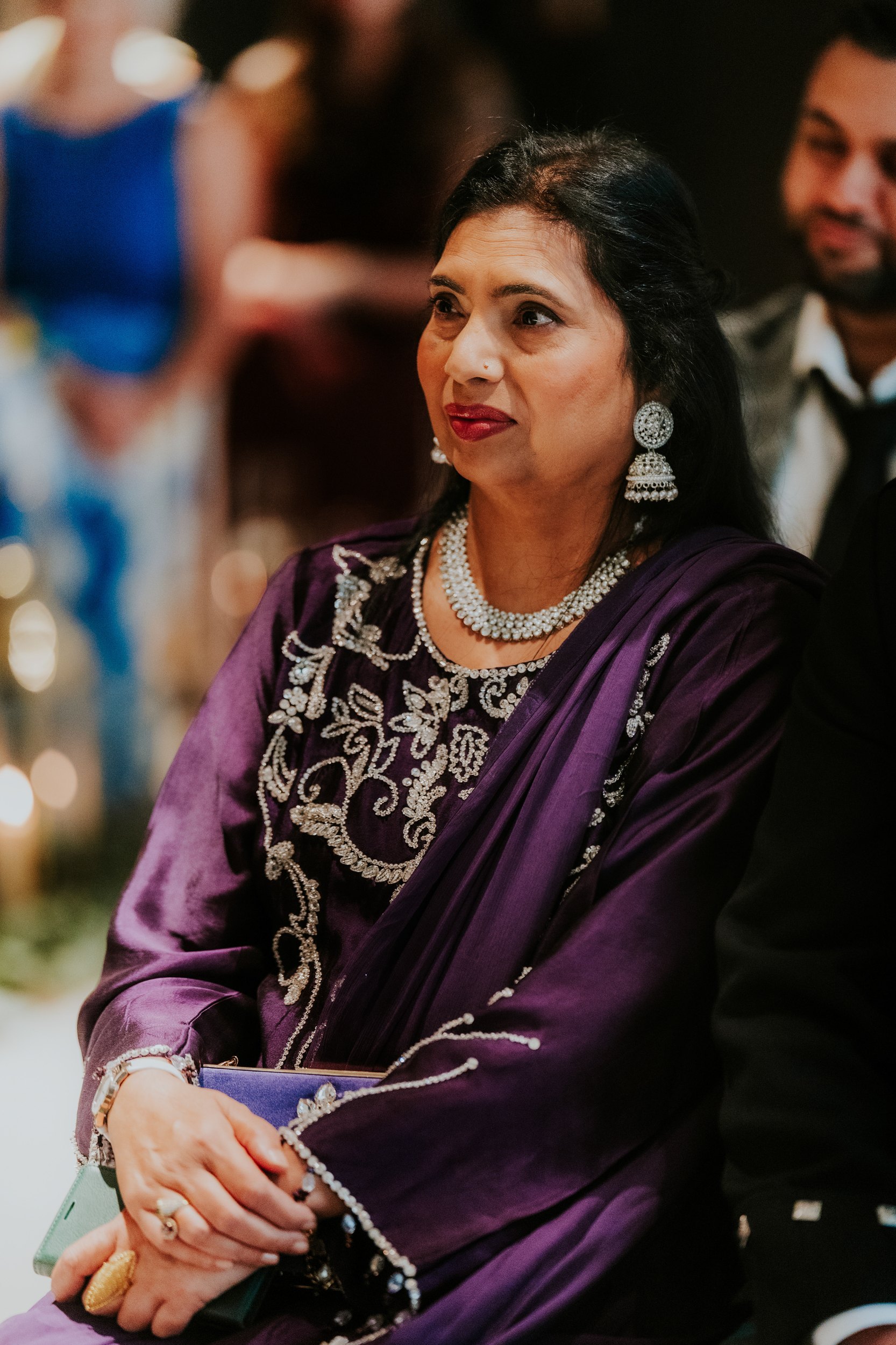 Woman in a purple embroidered dress with silver necklace and statement earrings, seated and looking to the side.