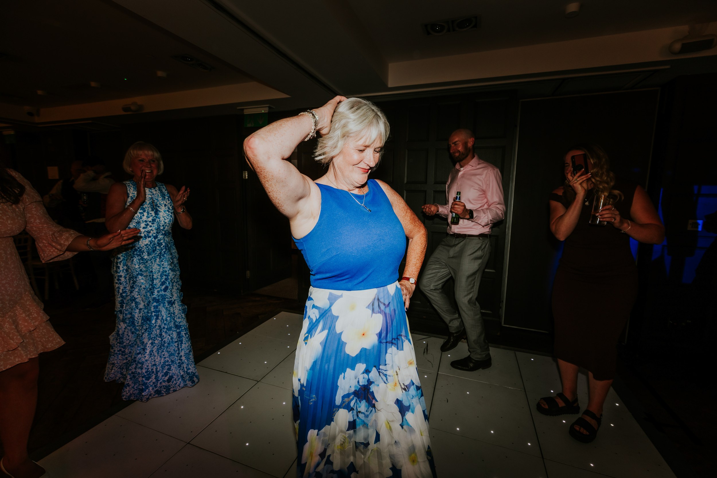 Woman in blue top and floral skirt dancing on a wedding dancefloor as guests clap and take photos.
