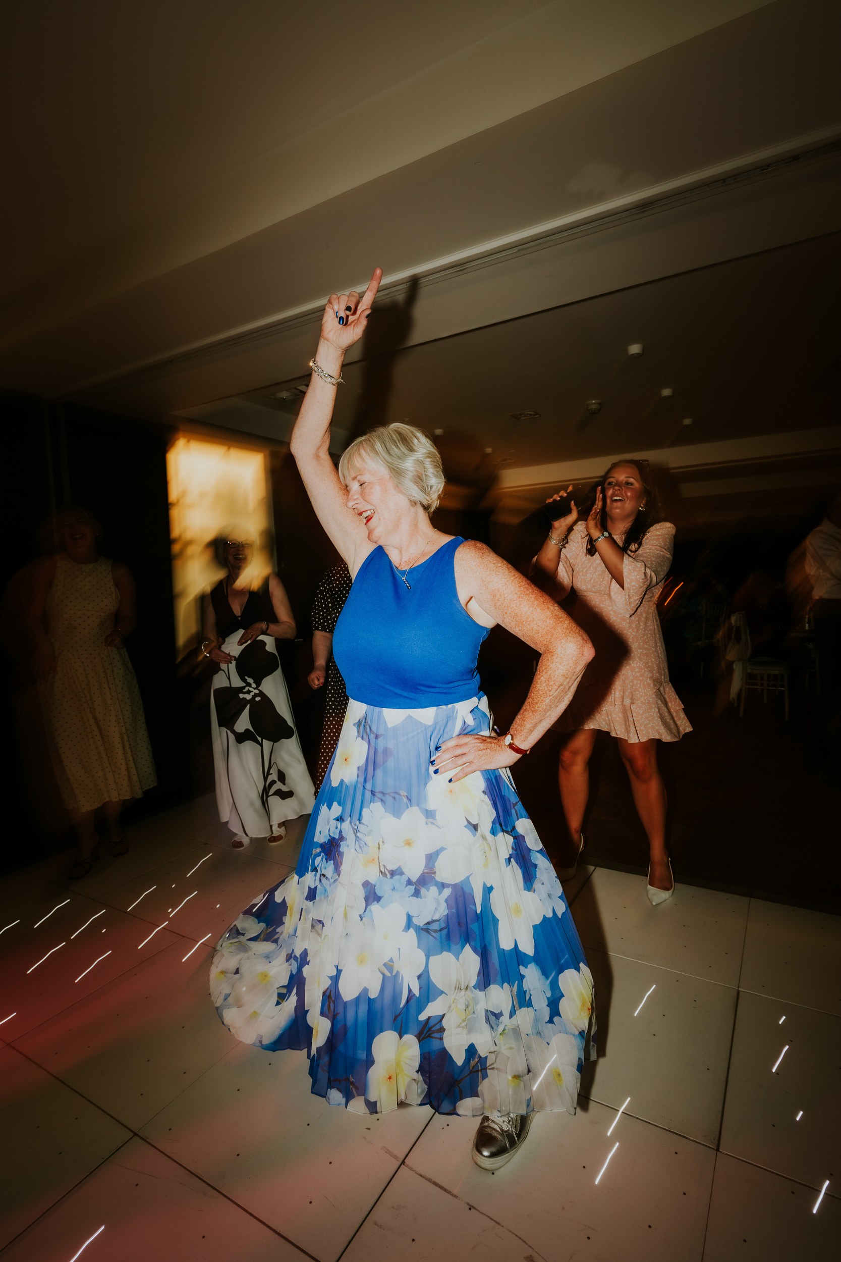 Woman in blue top and floral skirt dancing with arm raised on a wedding dancefloor, guests behind her.