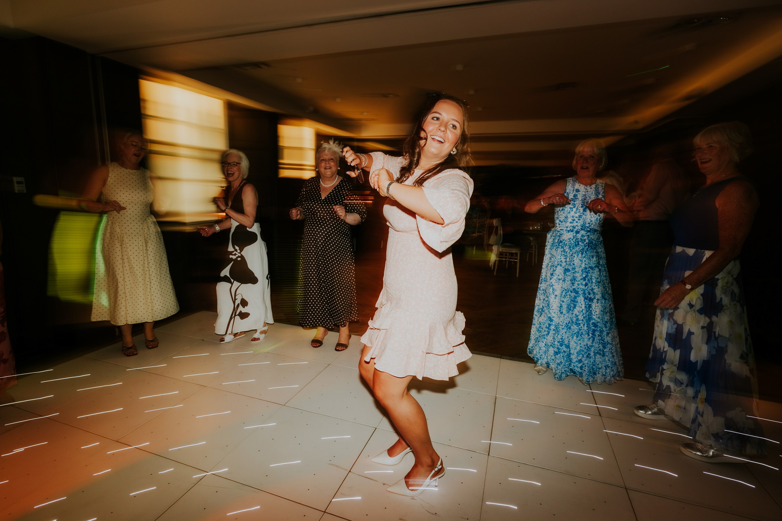 Young woman in pale pink dress dancing on a lit reception dancefloor with older female guests and light trails.