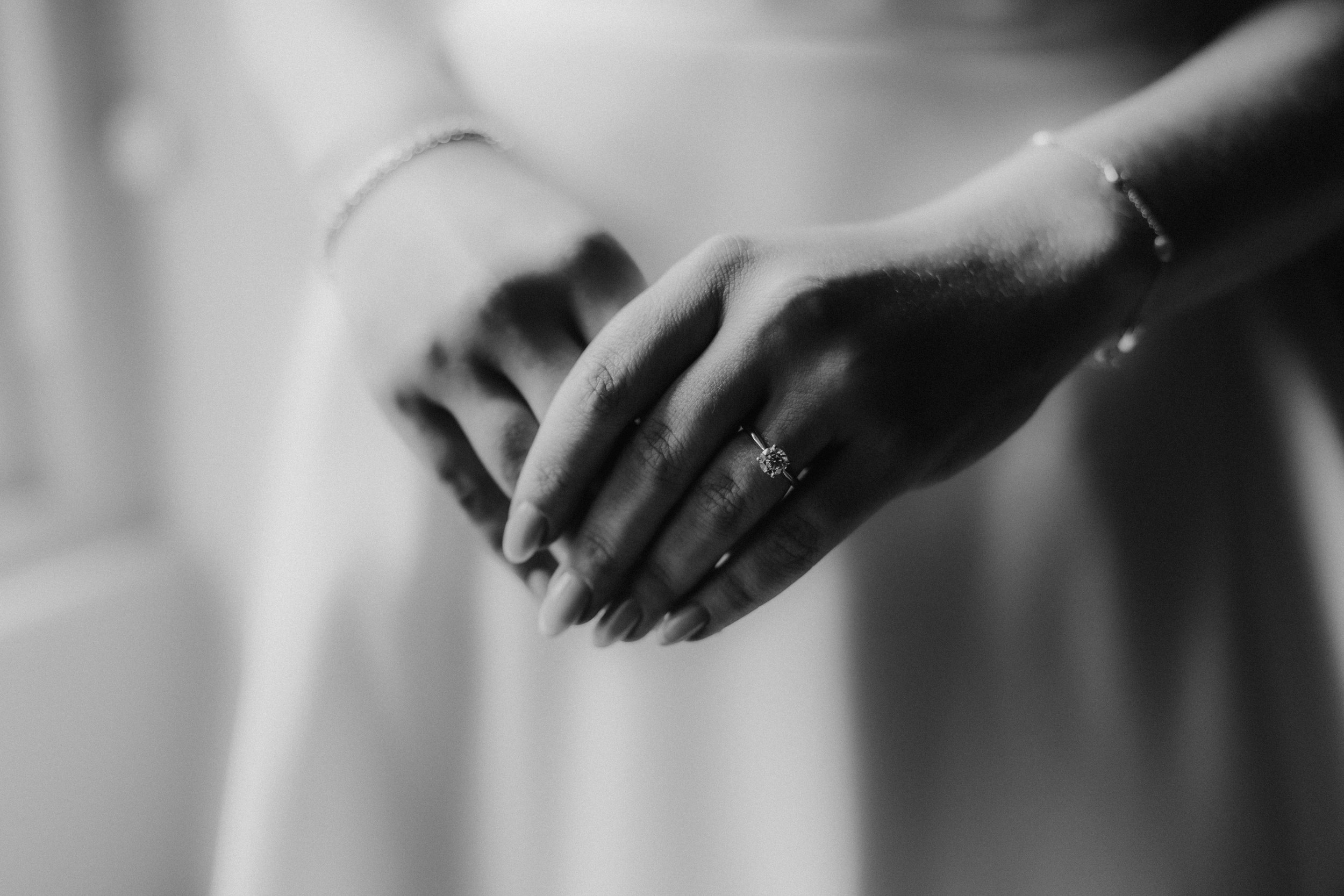 Black-and-white close-up of hands wearing a solitaire ring and bracelets against a blurred dress background.