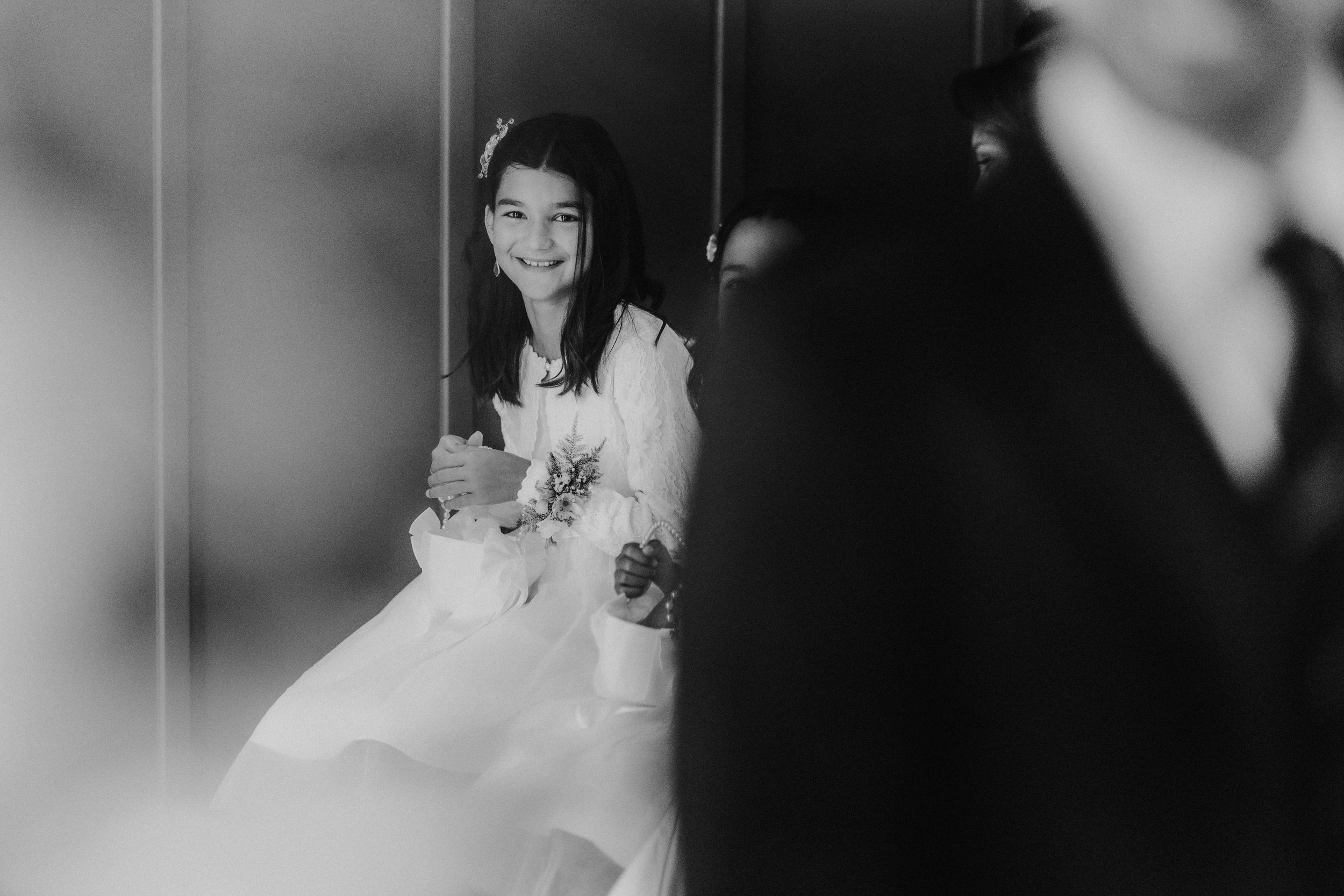 Black-and-white image of a smiling flower girl in a white dress holding a small bouquet or confetti basket.