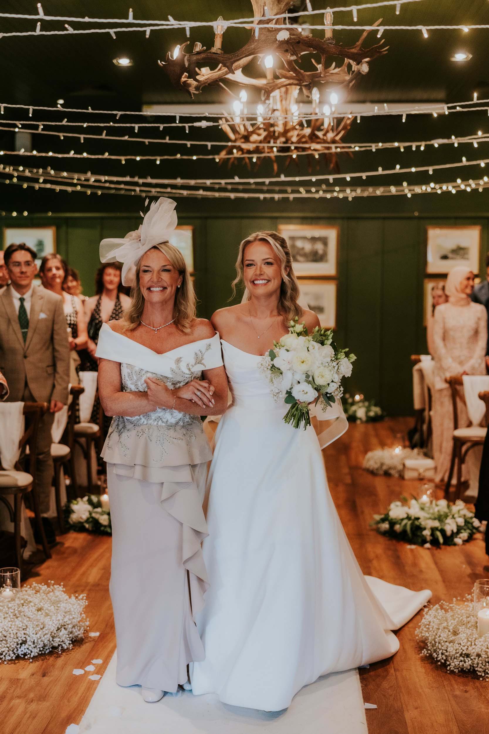 Bride Abigail walking down the aisle with her mother, smiling and holding a bouquet under string lights.