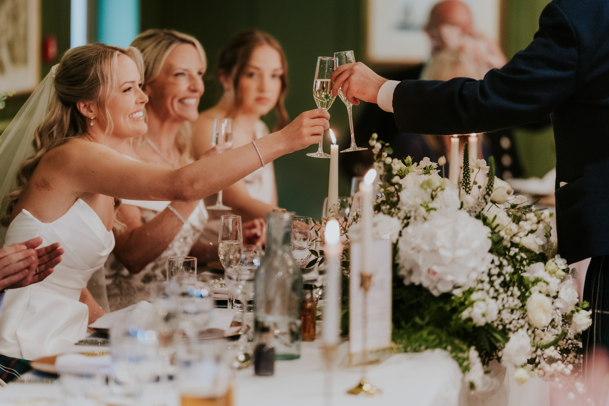 Bride and bridesmaids at the top table clinking champagne glasses over flowers and lit candles.
