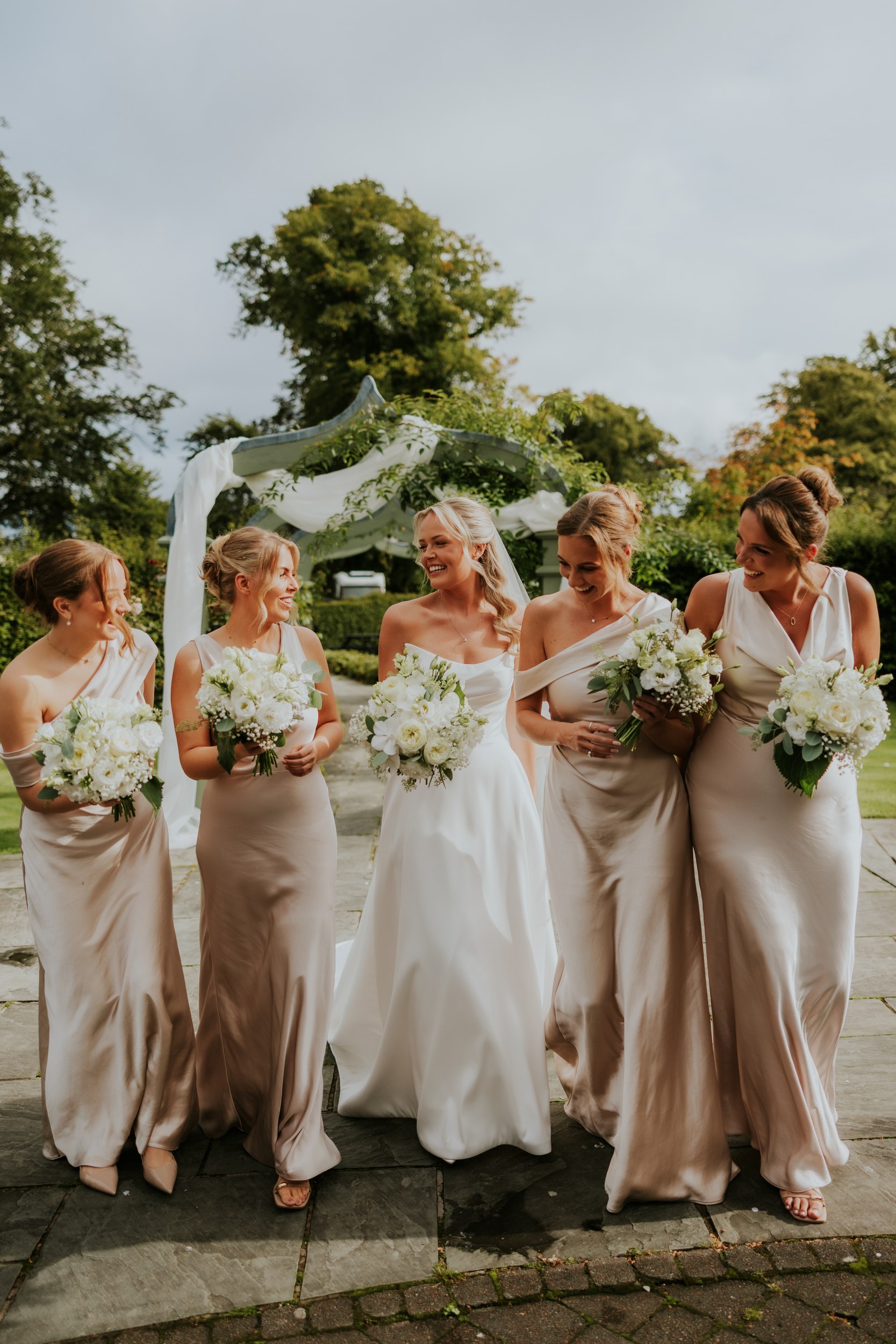Bride and four bridesmaids in cream dresses holding white bouquets, smiling together beneath a draped garden arch.