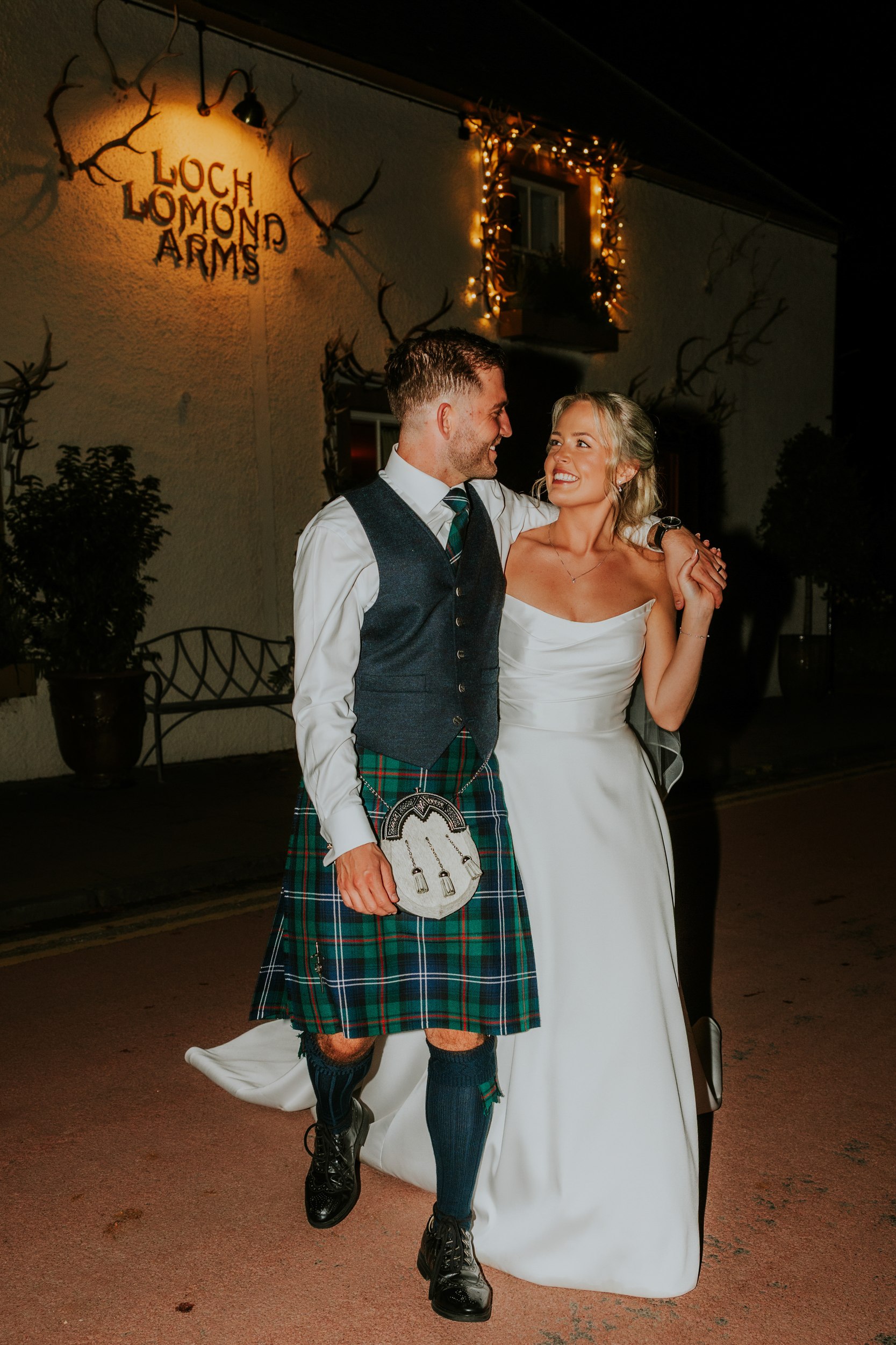 Bride and groom arm-in-arm outside the Loch Lomond Arms at night; groom in kilt, bride in white dress, fairy lights on the building.