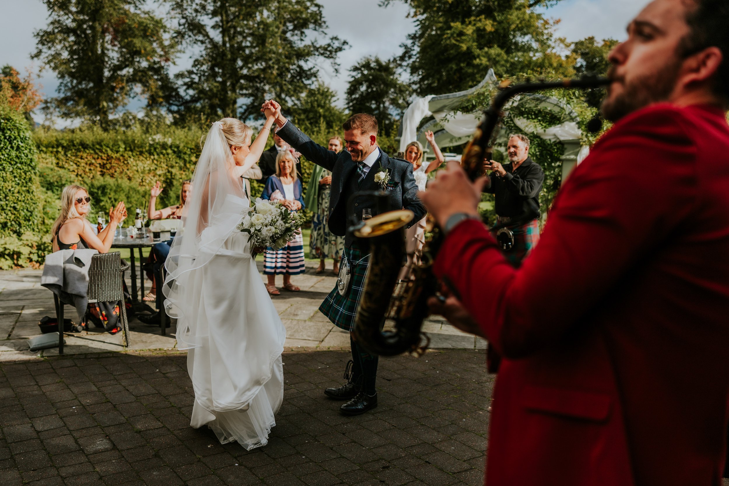 Bride and groom dancing outdoors with guests applauding and a saxophonist in the foreground.