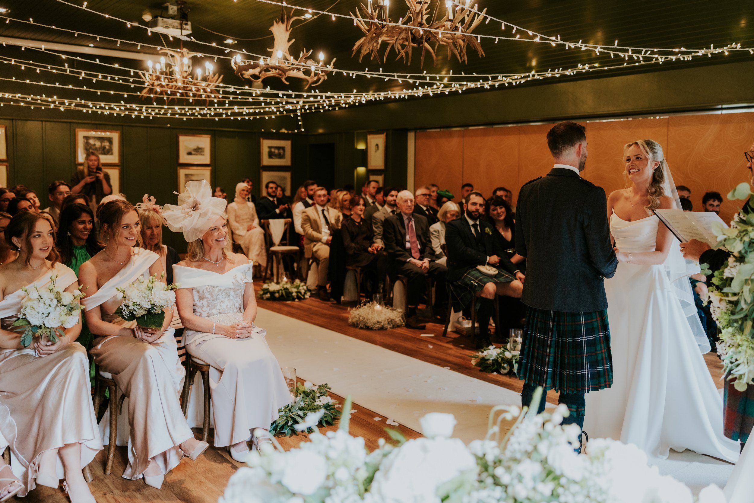 Bride and groom face each other at an indoor ceremony as bridesmaids and guests watch under fairy lights.