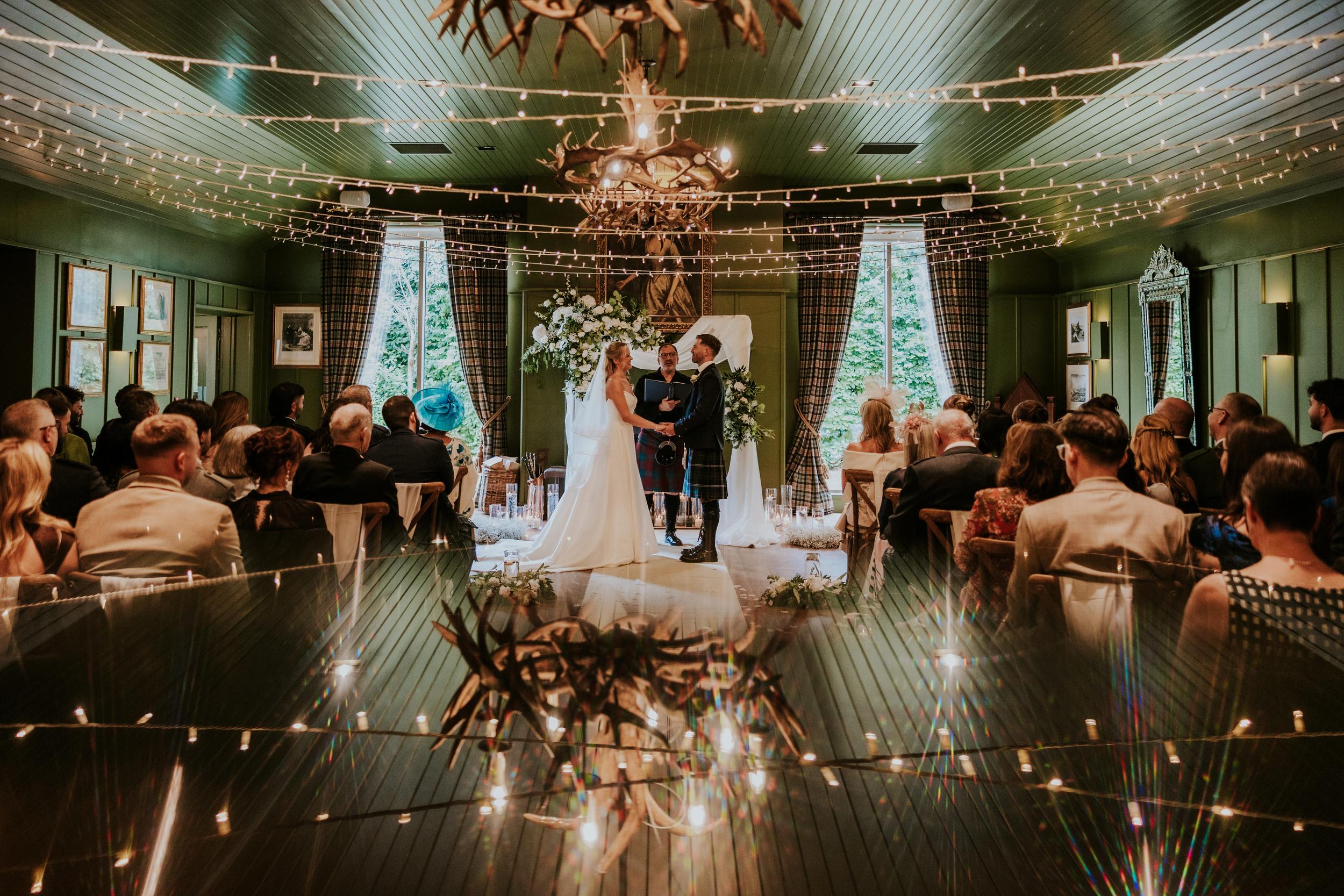 Bride and groom hold hands at an indoor ceremony altar under string lights while guests look on.