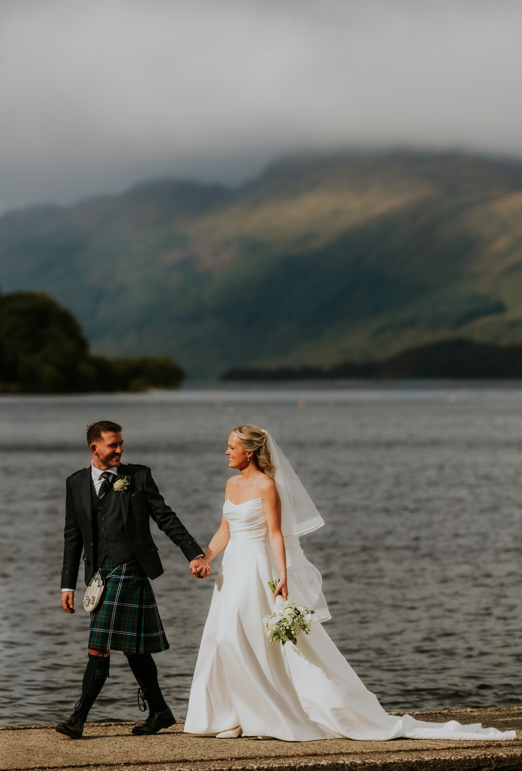 Bride and groom holding hands and walking beside a loch; bride in white dress with veil and bouquet, groom in a kilt.