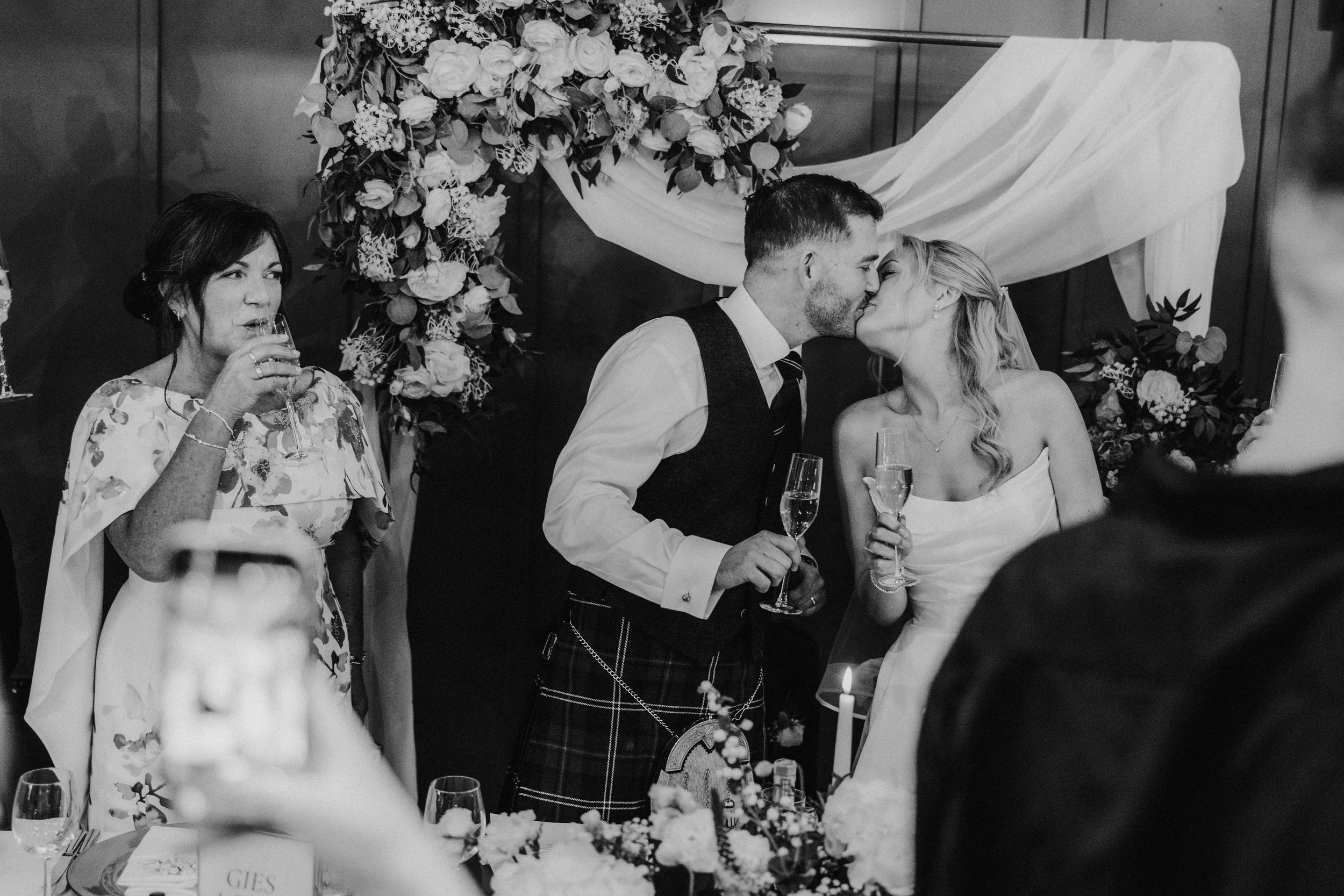 Bride and groom kiss at the top table during the reception as guests raise champagne; a woman at the table sips her drink.