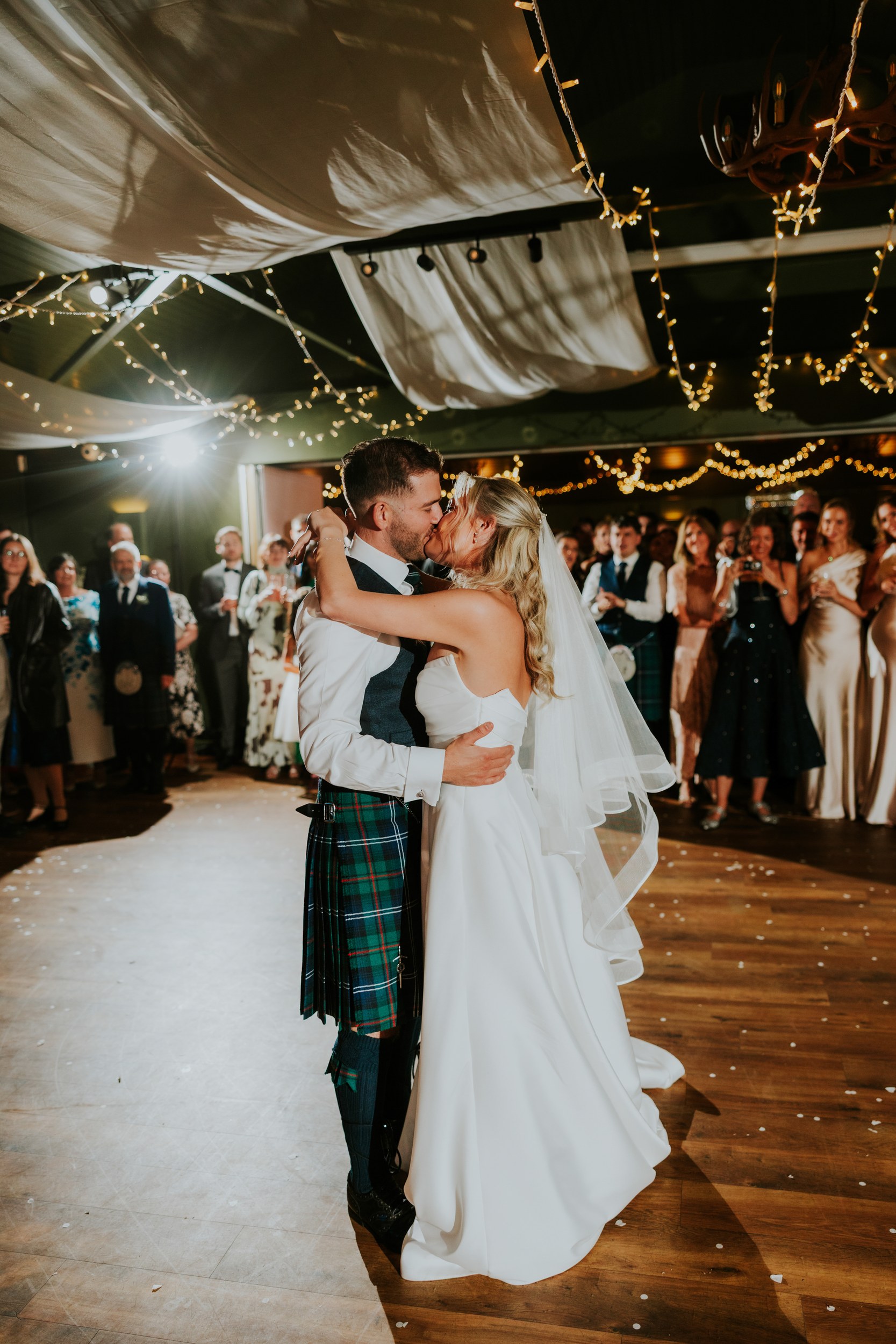 Bride and groom kiss during their first dance with guests watching under fairy lights.