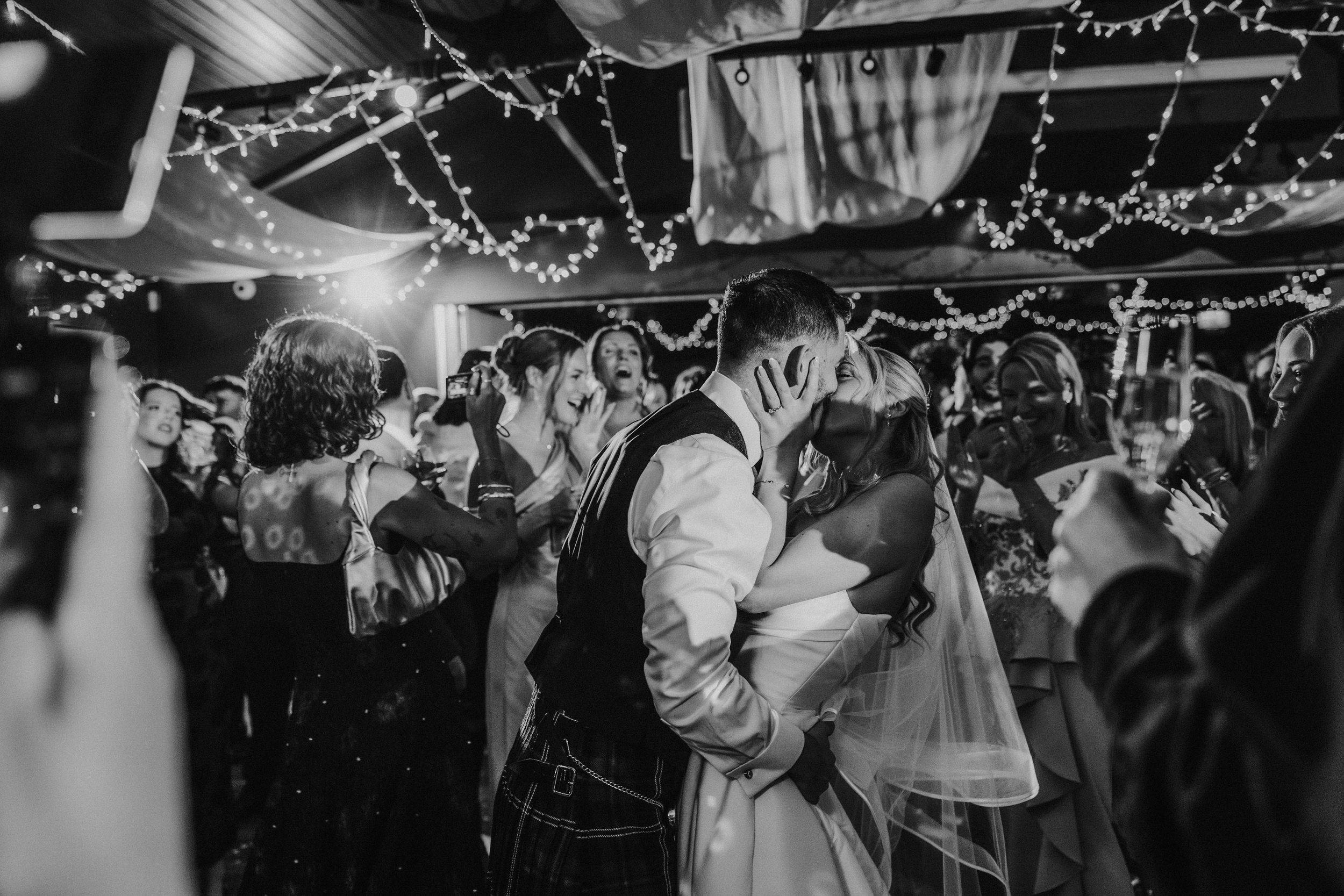 Bride and groom kiss on a crowded dancefloor under fairy lights as guests cheer and film on phones.