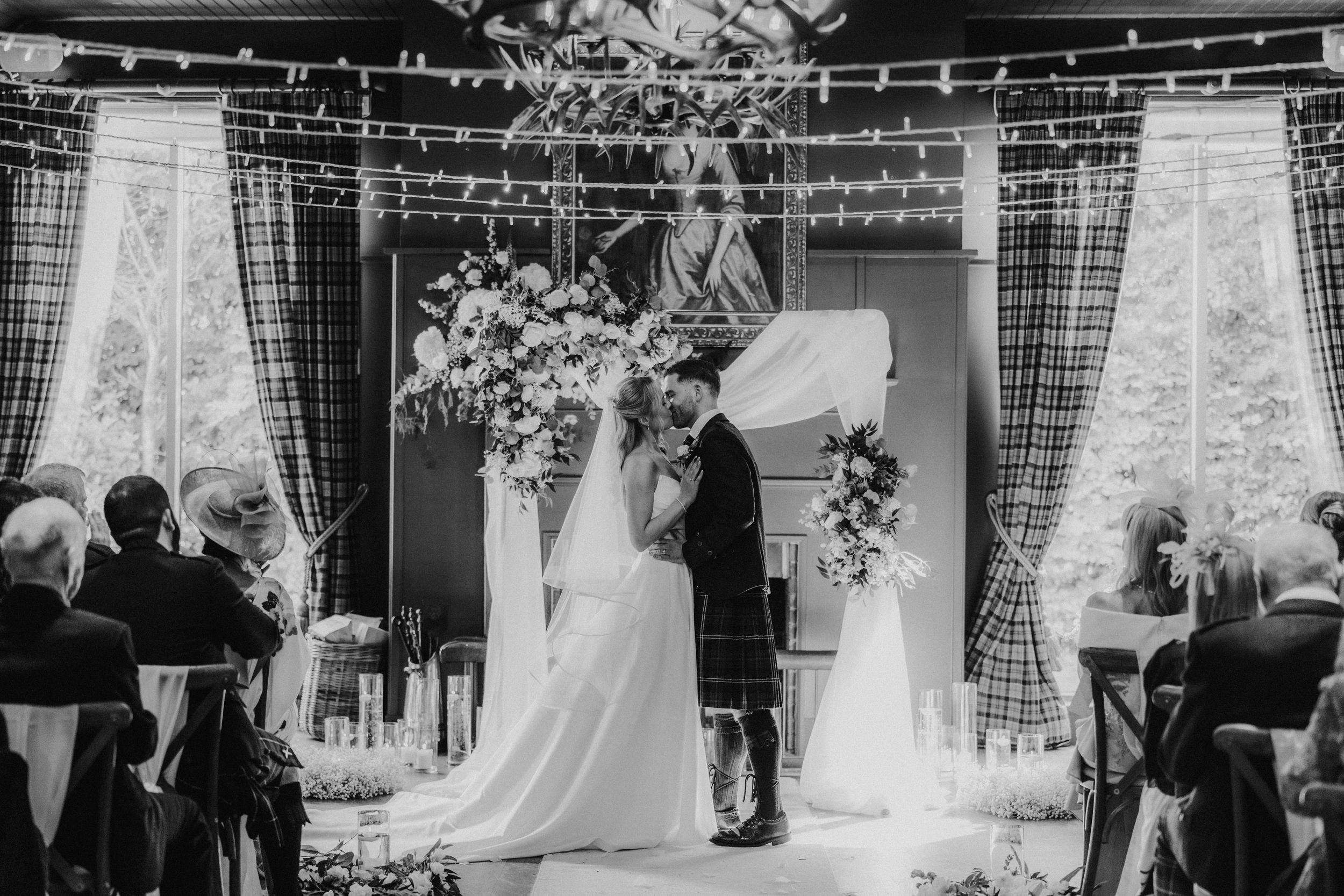 Bride and groom kiss under a floral arch inside a ceremony room with seated guests either side.