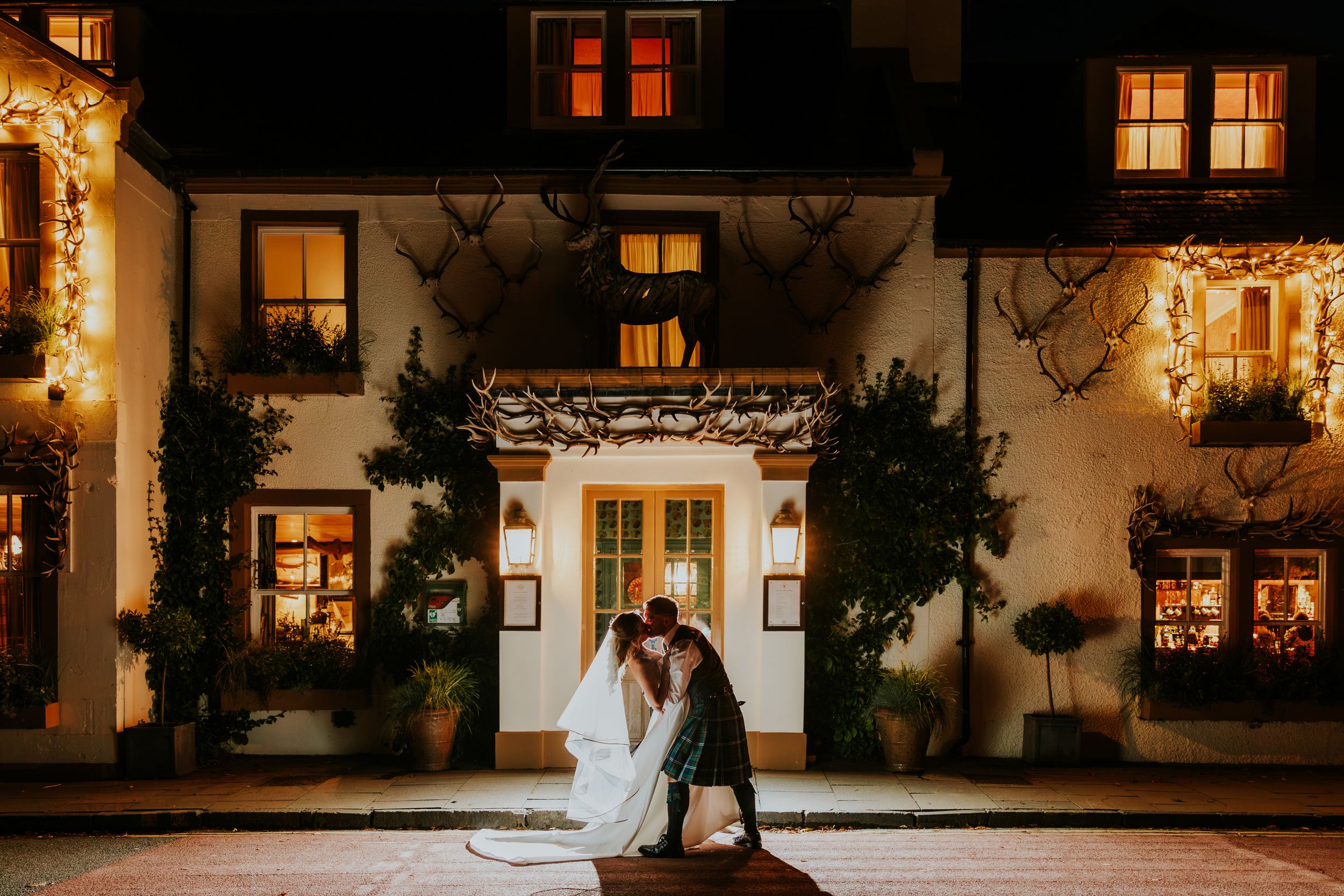 Bride and groom kissing outside the Loch Lomond Arms Hotel entrance at night with lit windows and antler decorations.