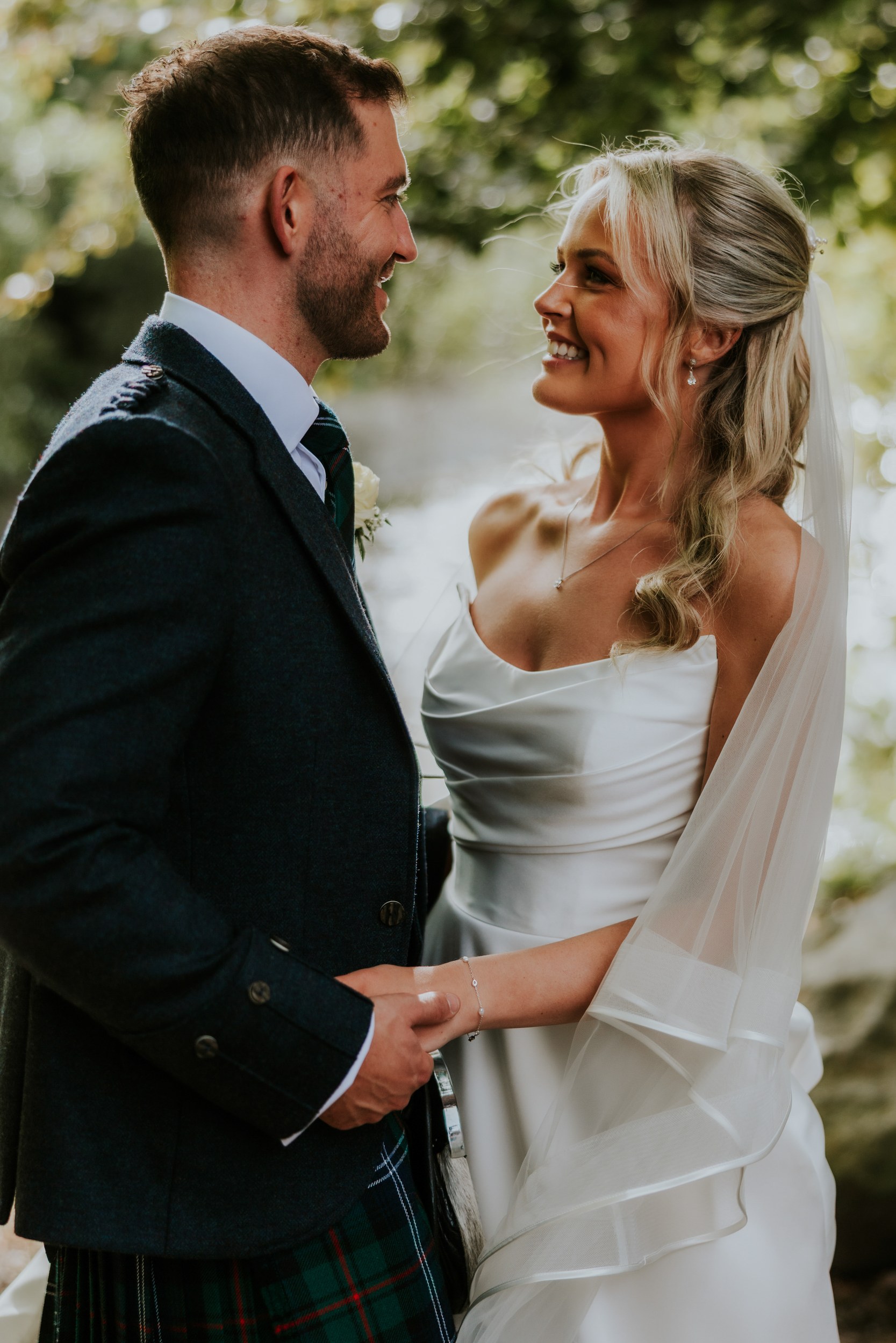 Bride and groom smile at each other holding hands; bride in strapless dress and veil, groom in jacket and tartan kilt, leafy background.