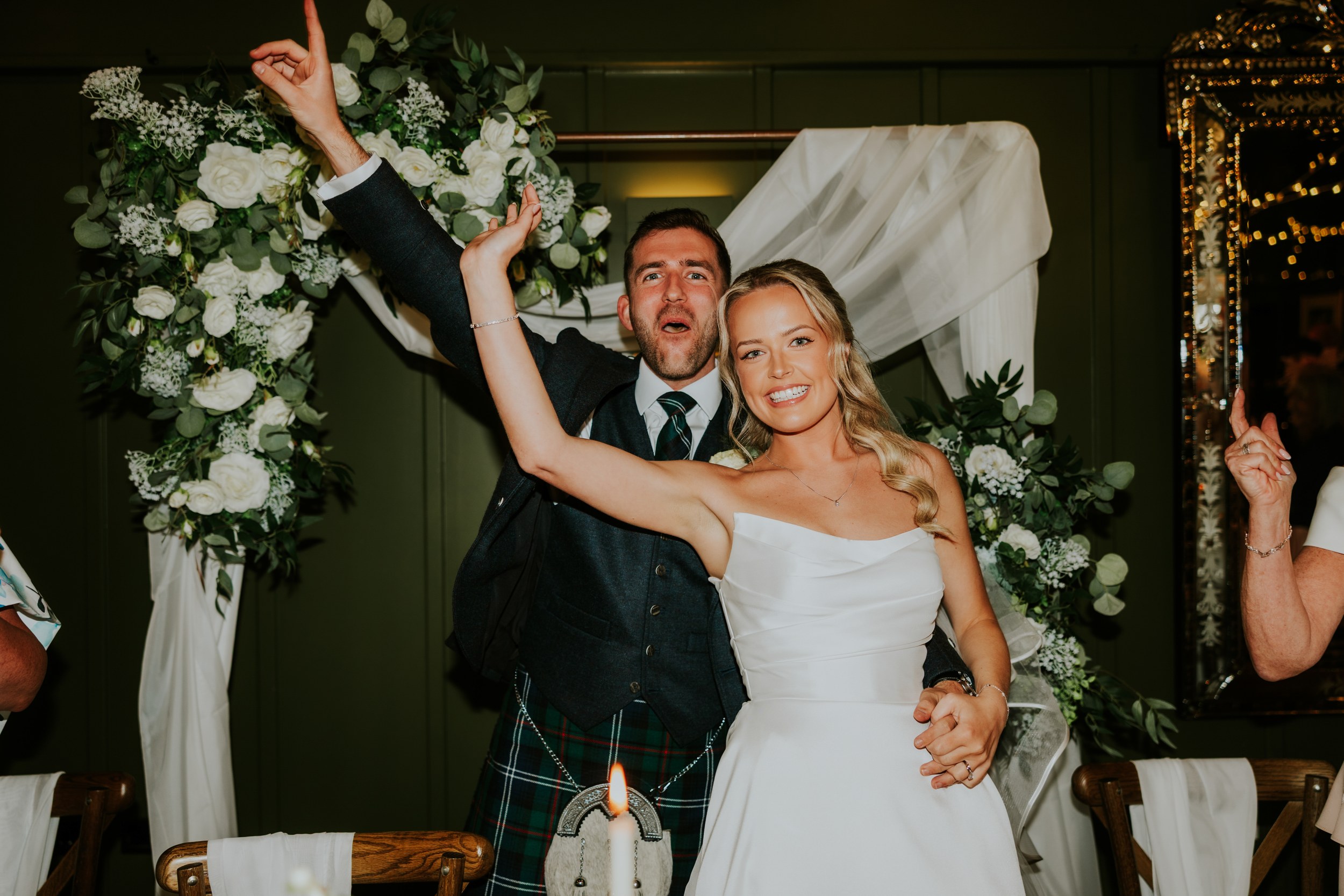 Bride and groom smiling with arms raised in front of a floral arch and draped fabric, candlelit table visible.