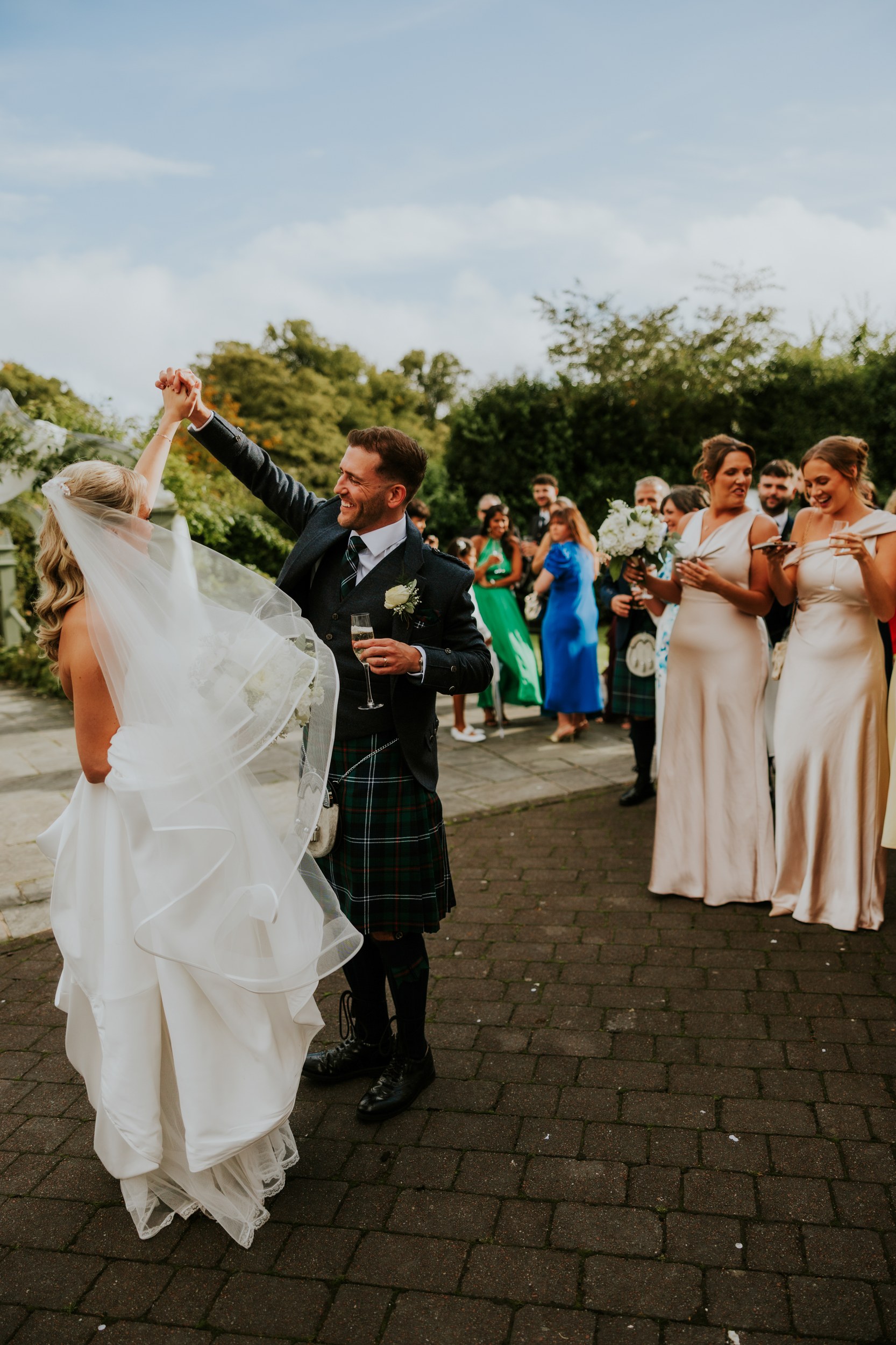 Bride and groom twirling outdoors as guests and bridesmaids watch, some holding drinks.