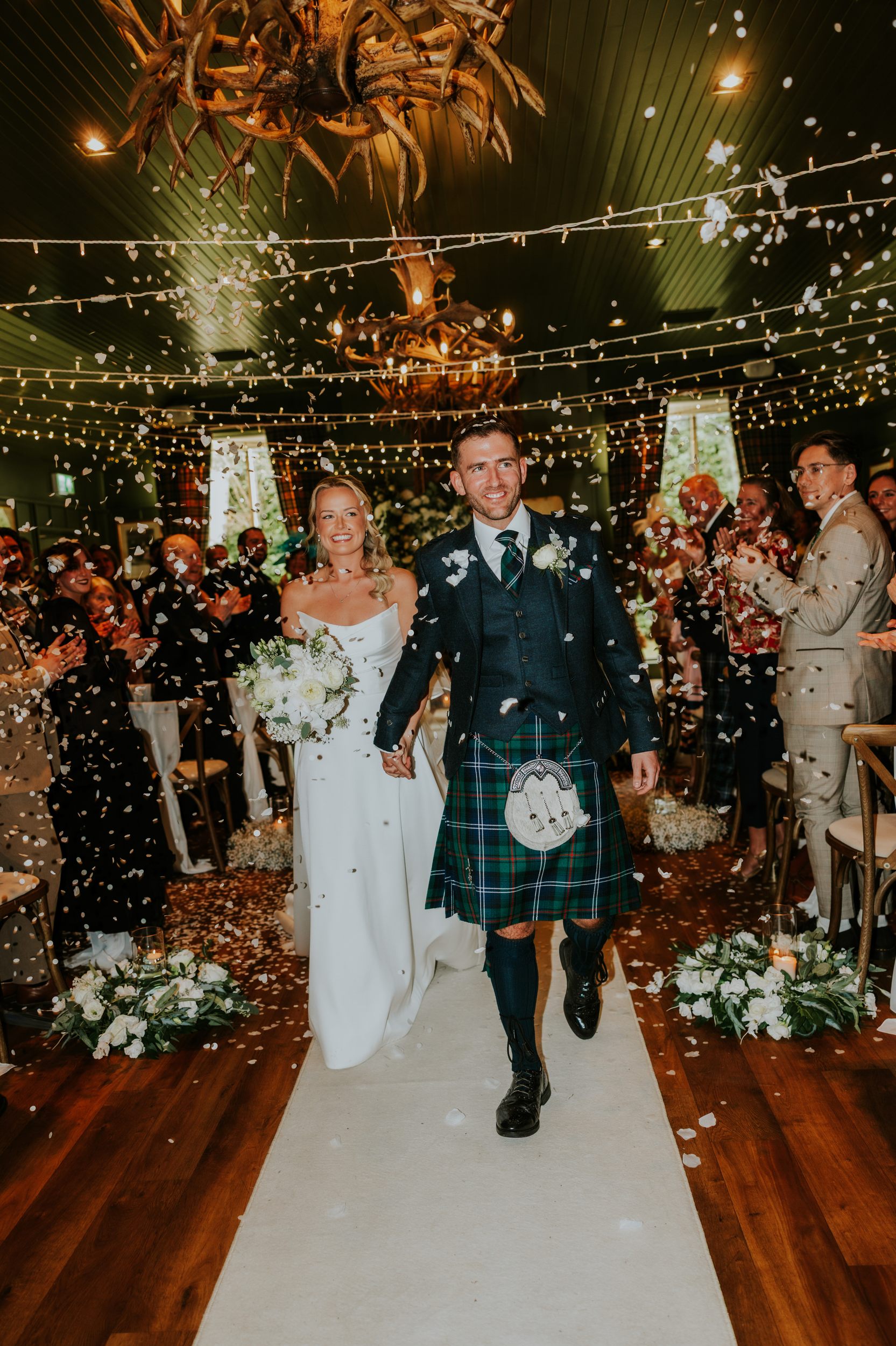 Bride and groom walk down the aisle as guests throw confetti; groom in tartan kilt, bride with white bouquet under fairy lights.