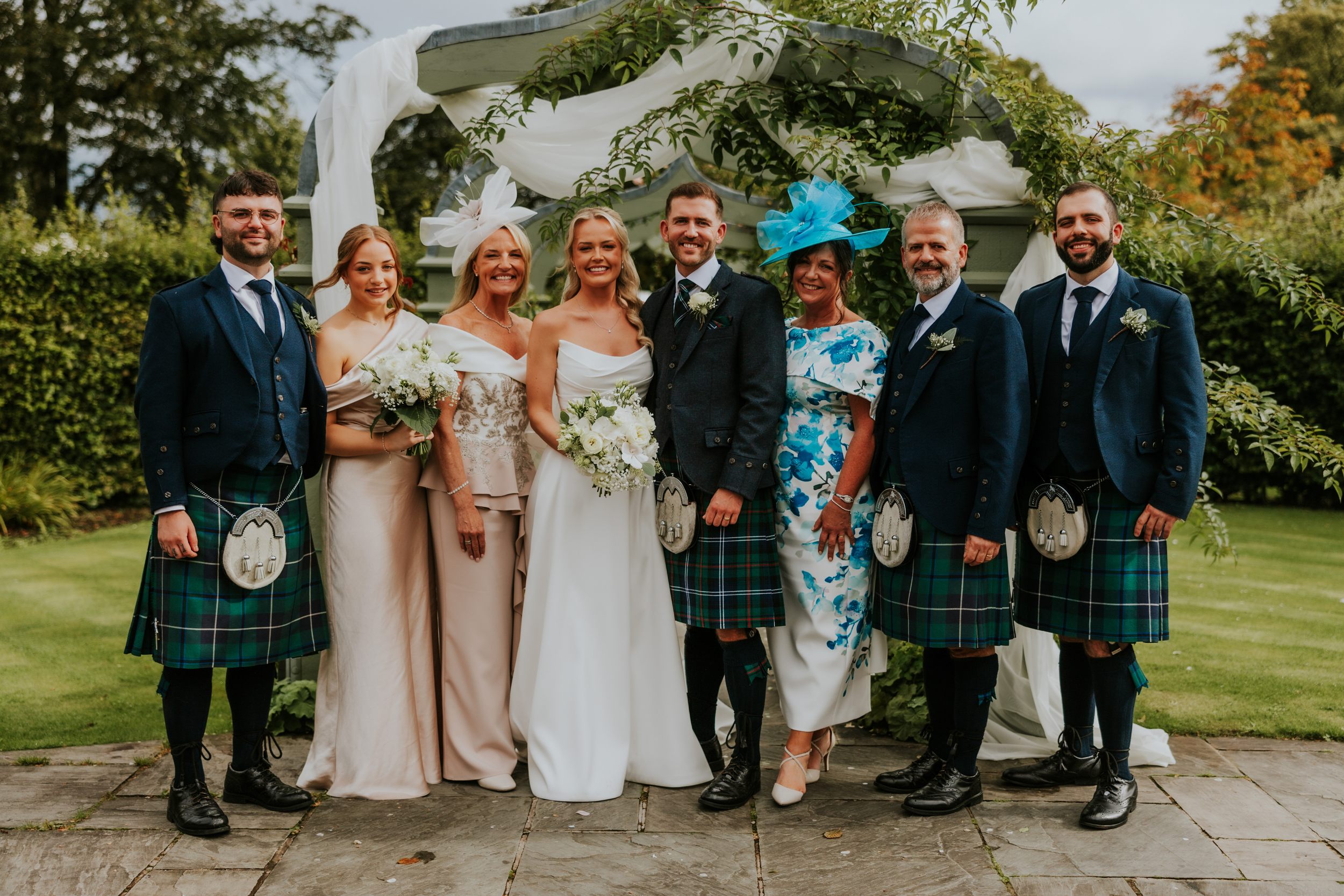 Bride and groom with bridesmaids and groomsmen in kilts posing under a draped garden arch, all smiling.
