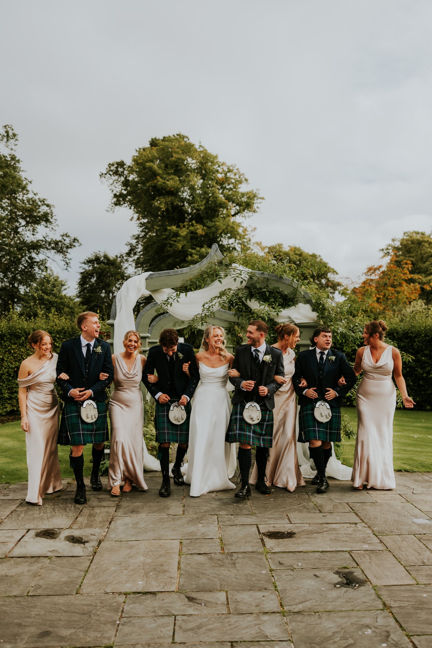 Bride and groom with bridesmaids and kilted groomsmen linked arm-in-arm beneath a draped garden arbor.
