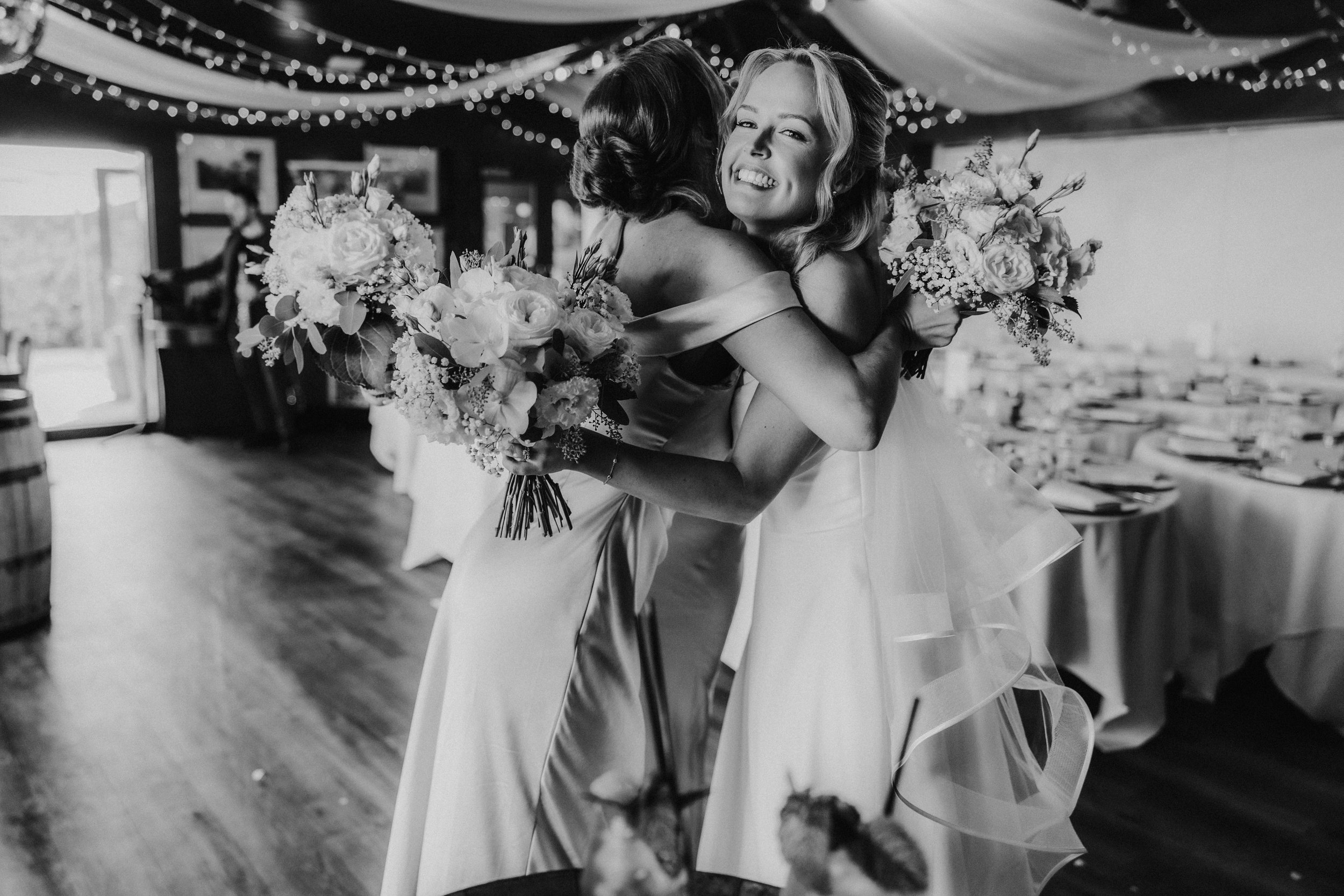 Bride hugging a bridesmaid in the decorated reception room, both holding bouquets.