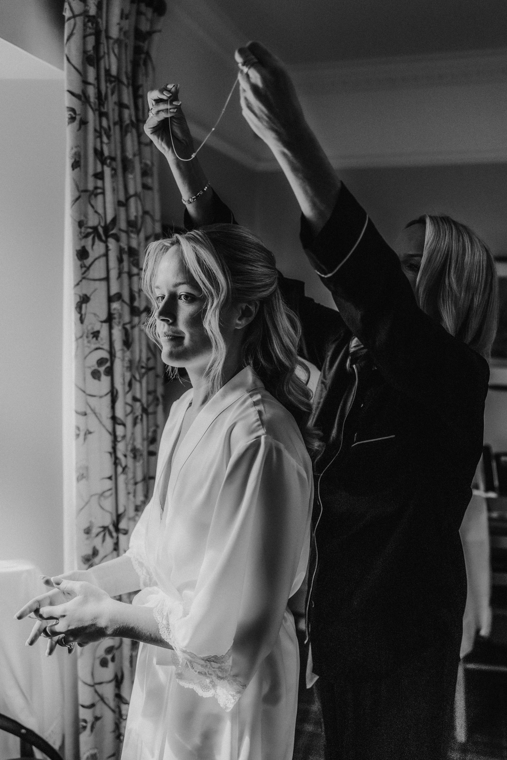 Bride in a robe having a necklace fastened by a woman during morning preparations.