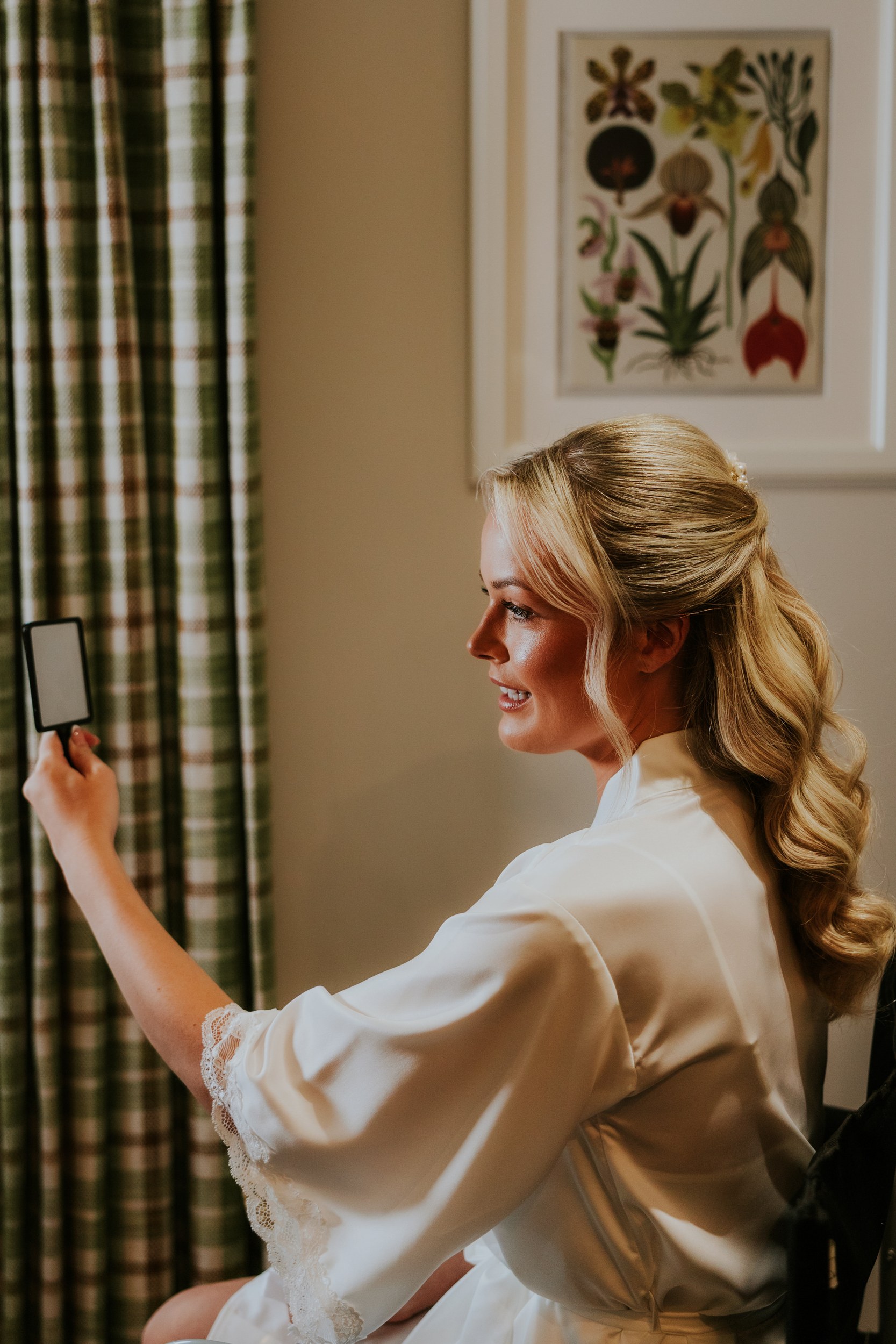 Bride in a satin robe checking her makeup in a small hand mirror in a hotel room.