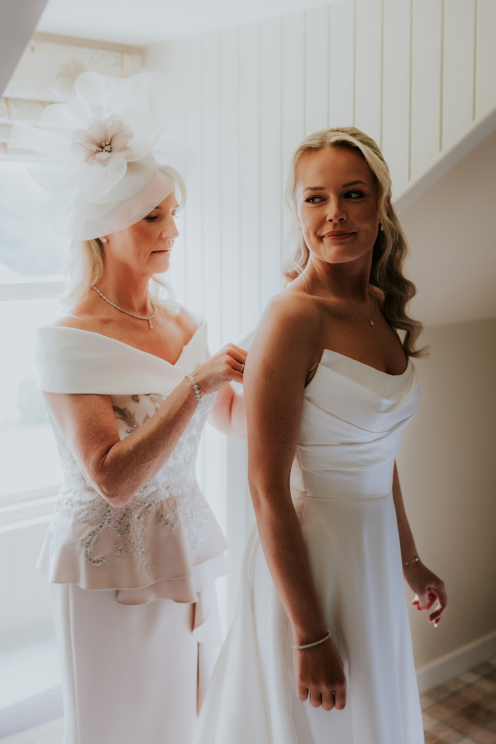 Bride in a strapless dress looks over her shoulder while an older woman fastens the back of her gown by a window.