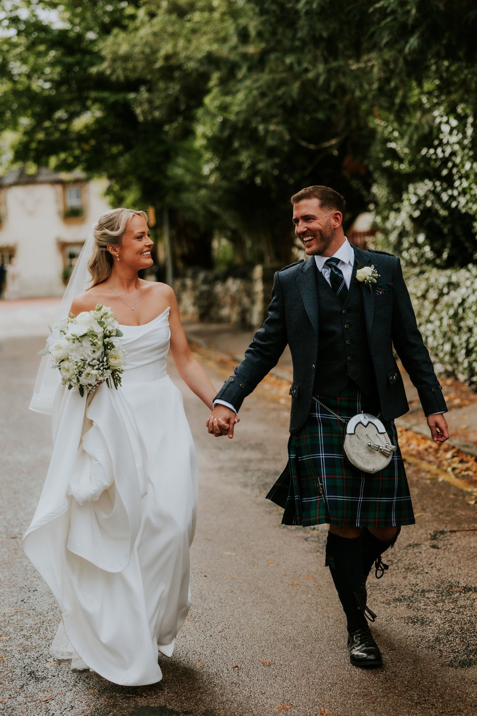 Bride in a white dress holding a bouquet and groom in a kilt walk hand-in-hand, smiling on a tree-lined street.
