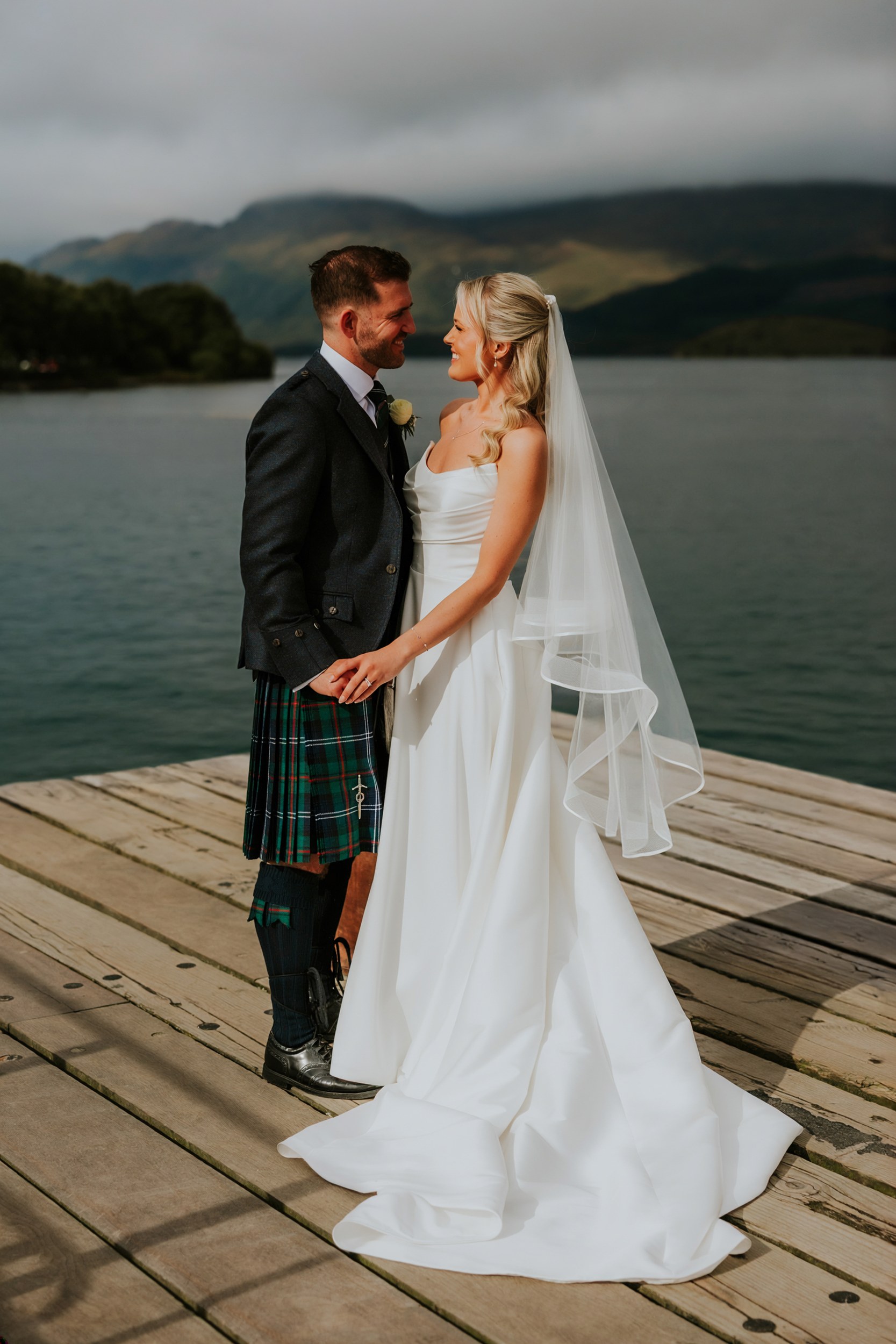 Bride in a white gown and veil and groom in a kilt smiling and holding hands on a wooden jetty by a loch with hills behind.
