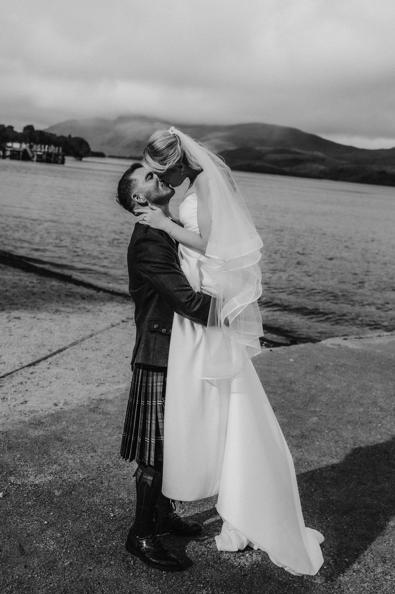 Bride in dress and veil lifted by groom in a kilt, kissing by the loch with hills in the background.