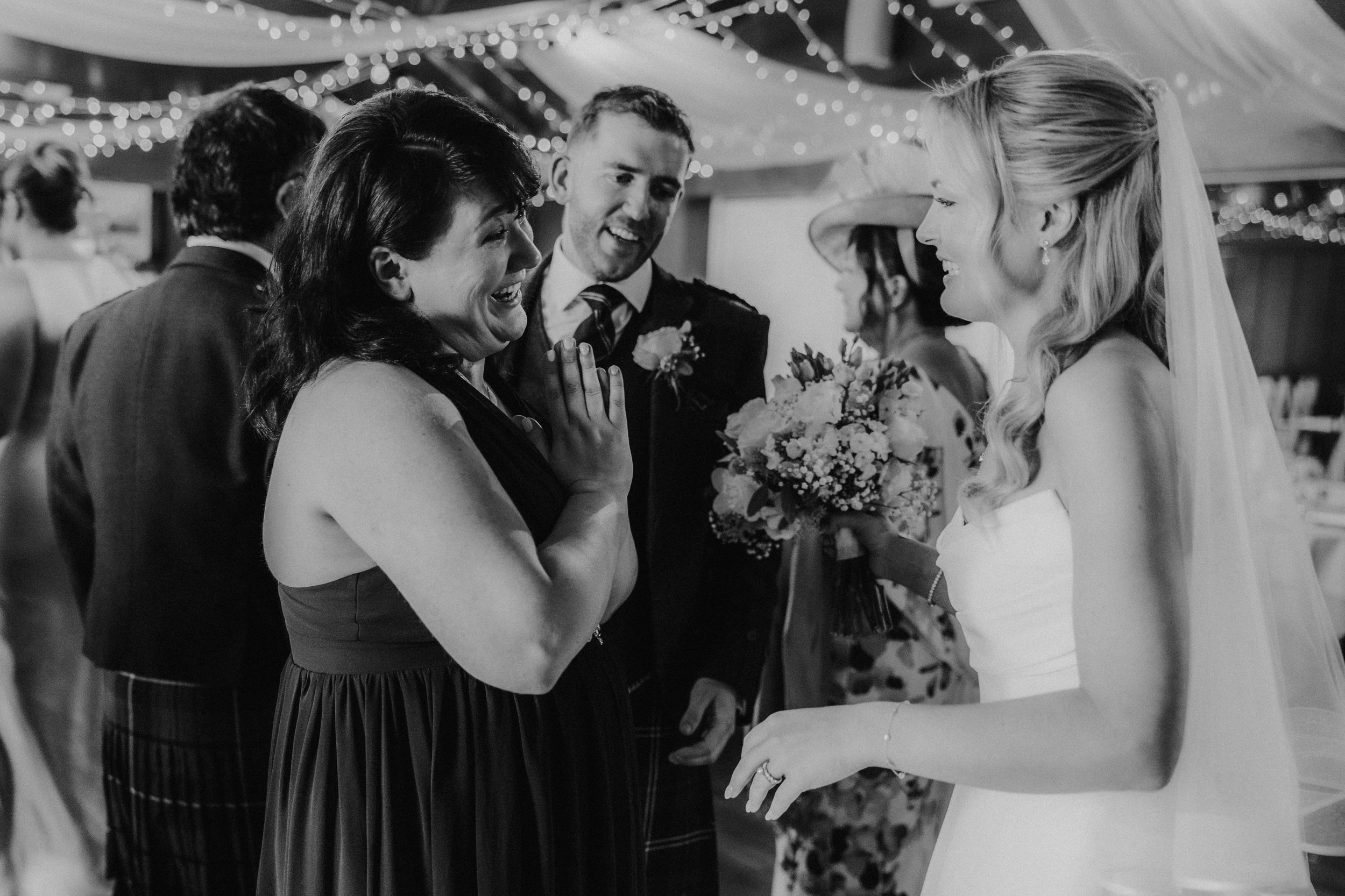 Bride in dress and veil smiling while chatting with delighted guests under fairy lights, a guest clasps her hands in surprise.