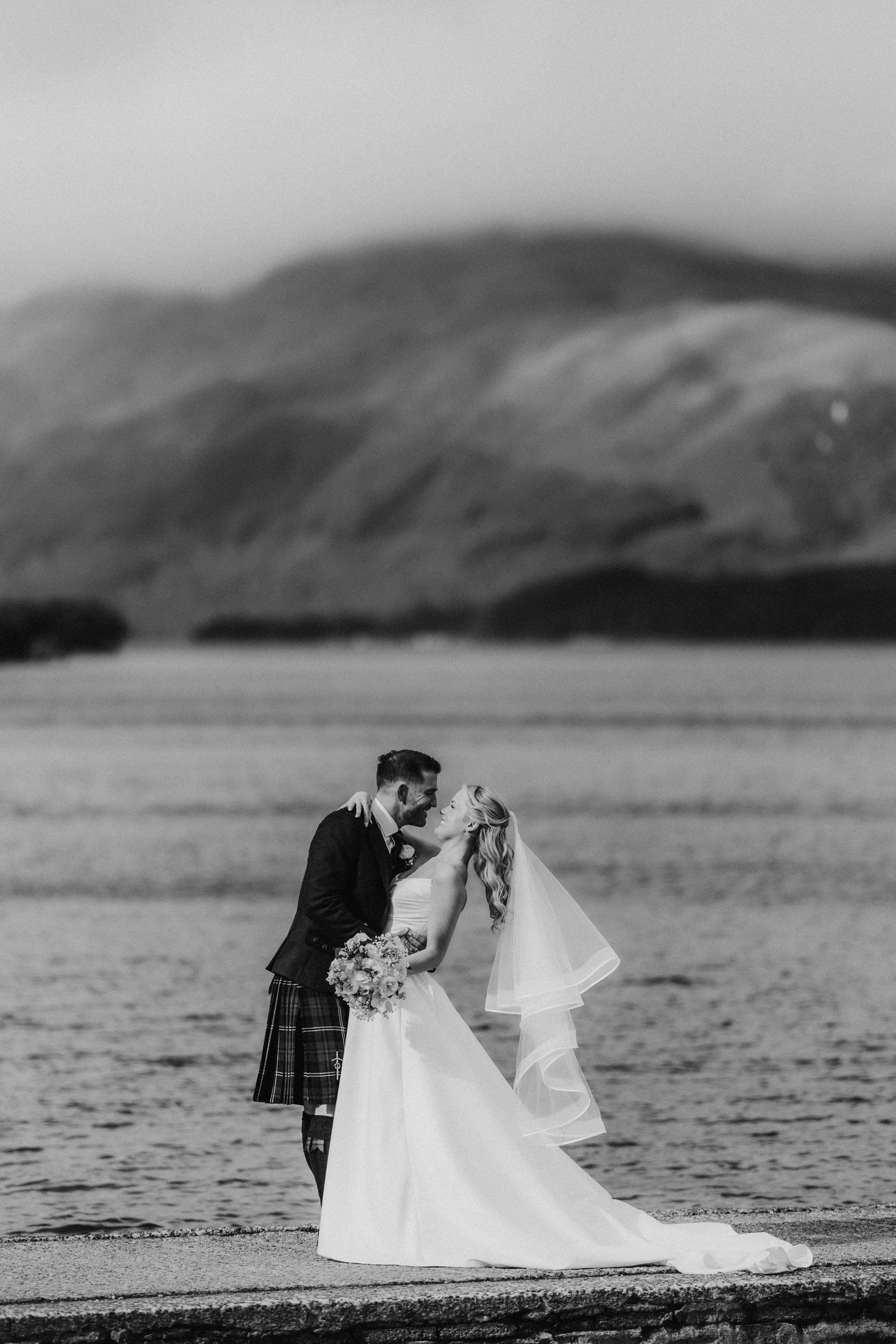 Bride in gown and veil and groom in kilt share a dip and kiss on a loch shore with hills behind them.