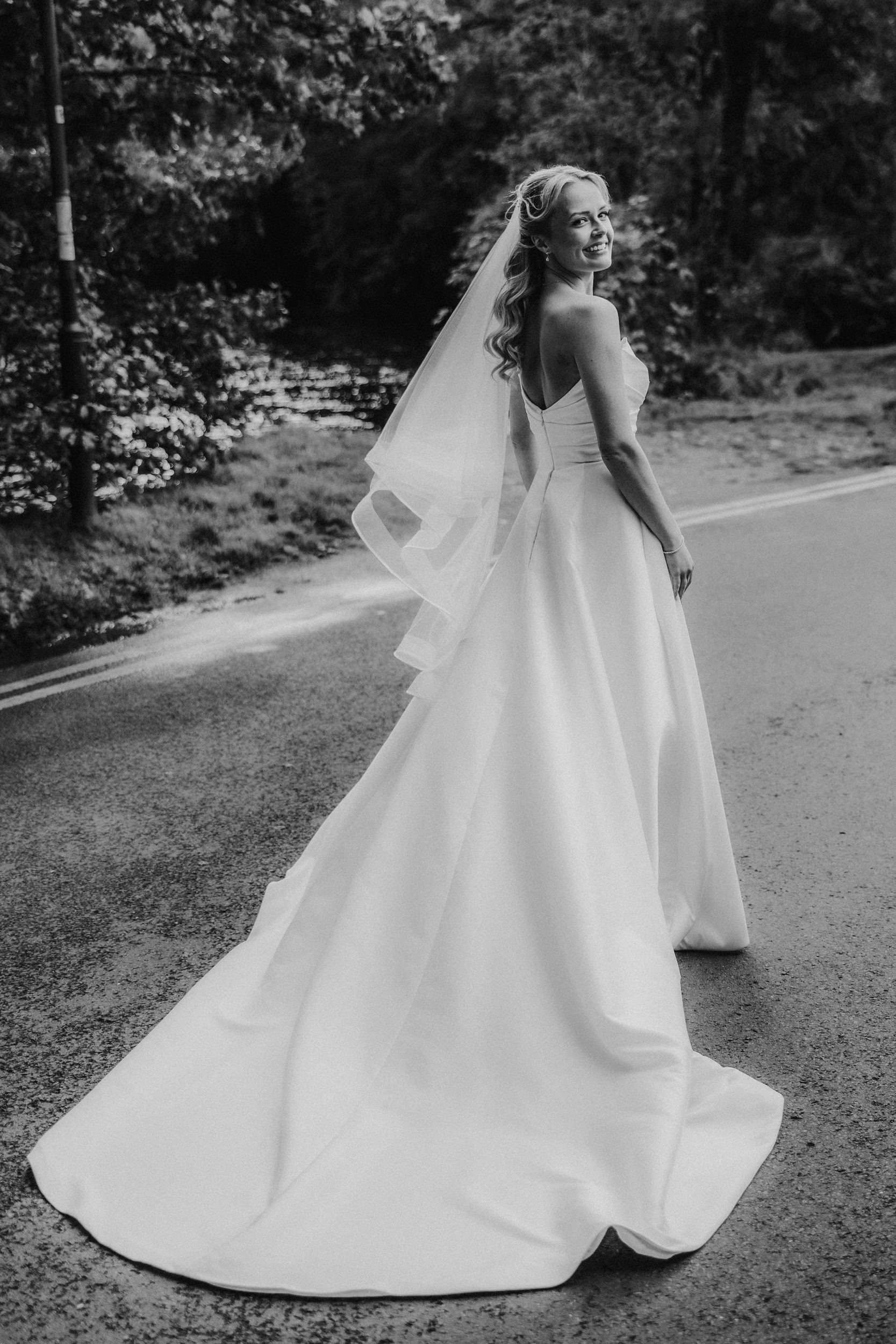 Bride in strapless dress and long veil stands on a road by trees and water, looking back and smiling.