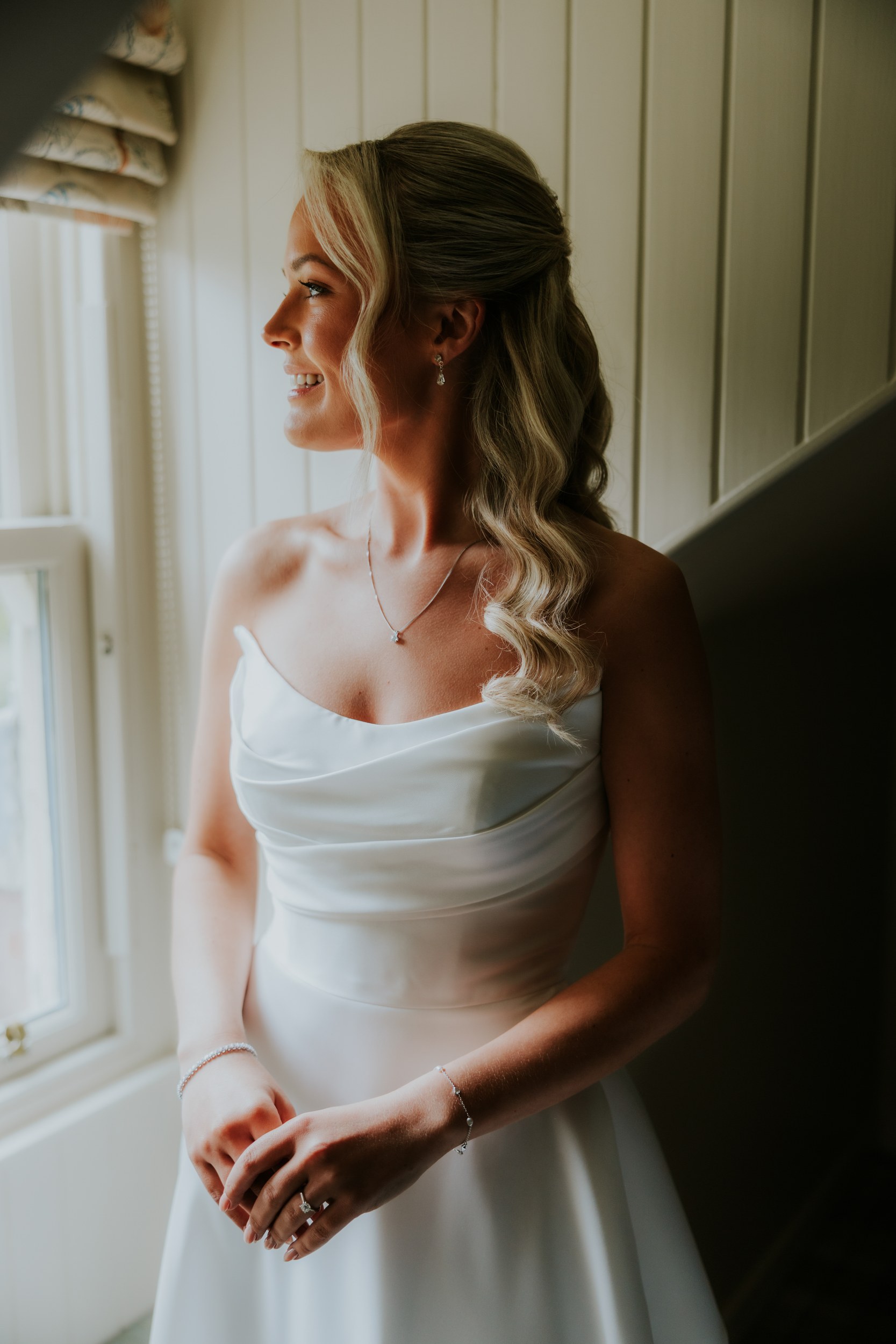 Bride in strapless dress smiling and looking out of a window, side-profile showing hair, necklace and hands with ring.