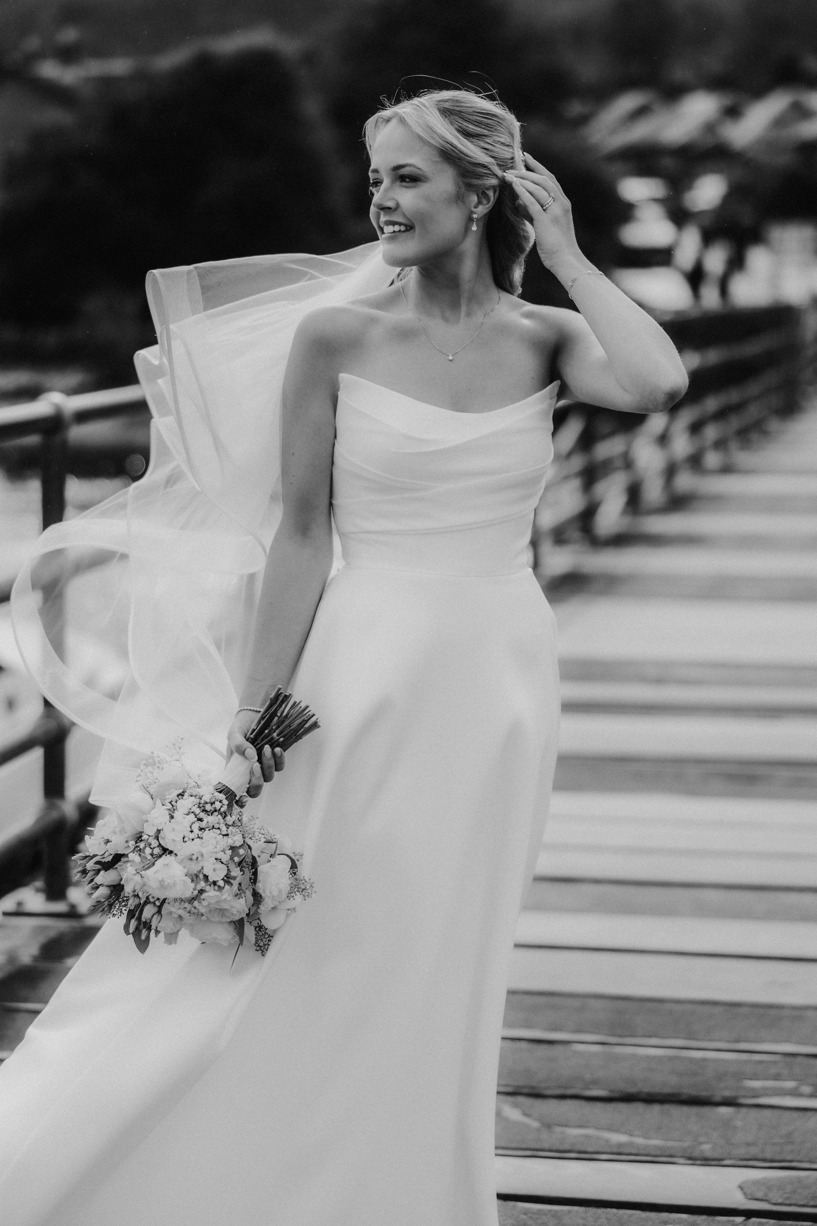 Bride in strapless gown adjusting her hair, holding bouquet with veil blowing on a wooden walkway by the water.