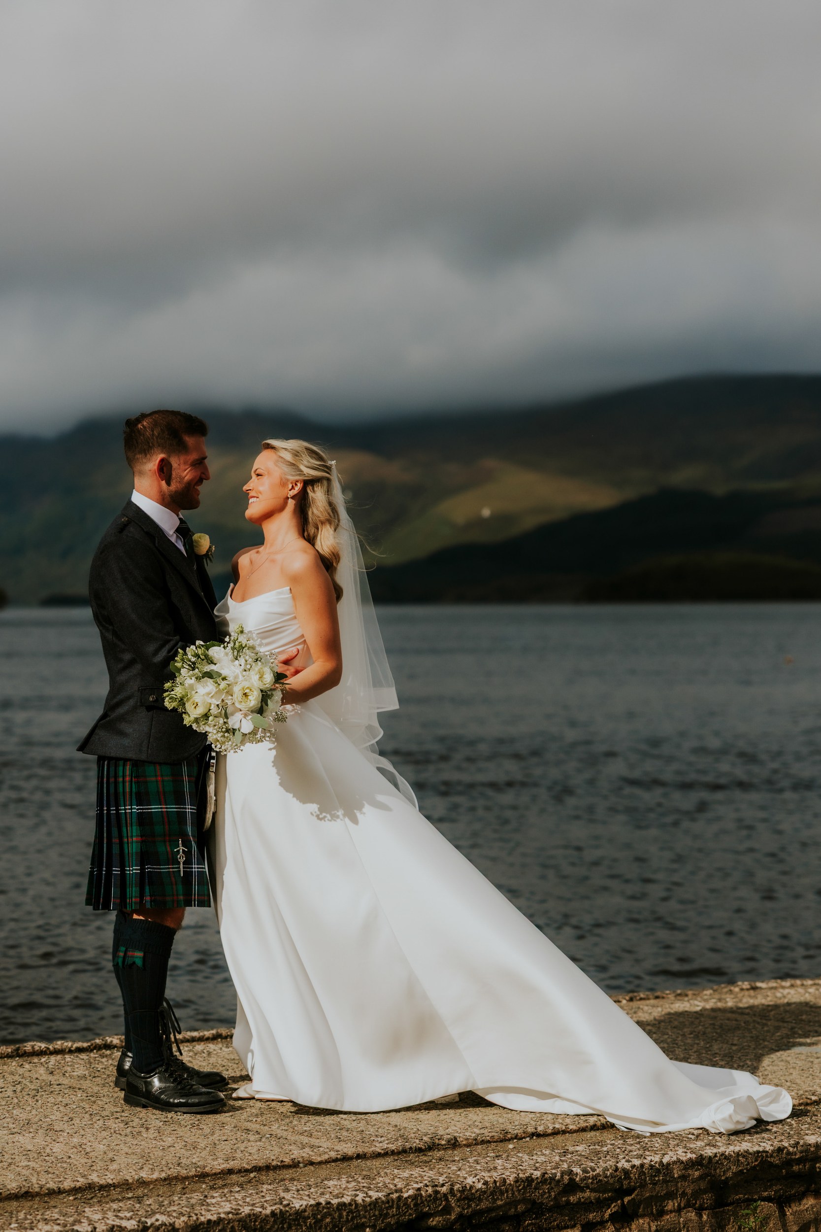 Bride in strapless gown and groom in kilt smiling by the loch with hills and cloudy sky behind.