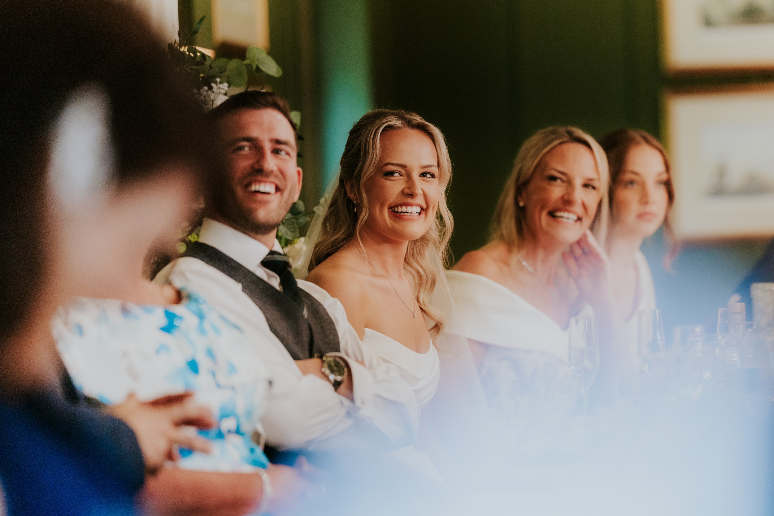 Bride in white and groom smiling with two female guests seated together at the wedding reception.