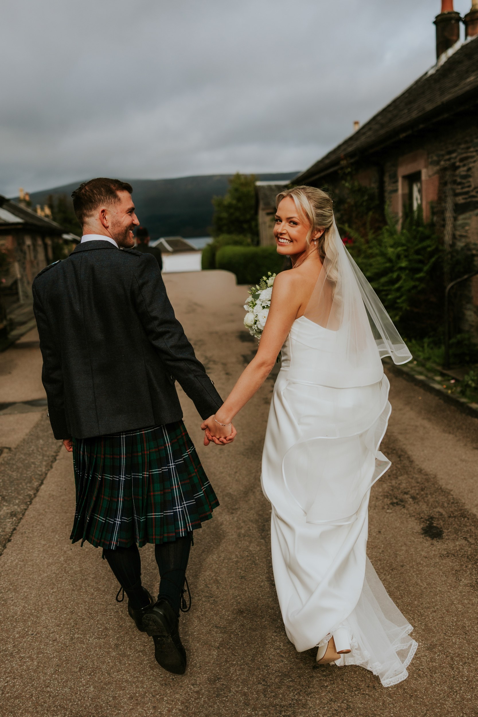 Bride in white dress and veil holding a bouquet looks back and smiles while holding hands with groom in a tartan kilt on a lane.