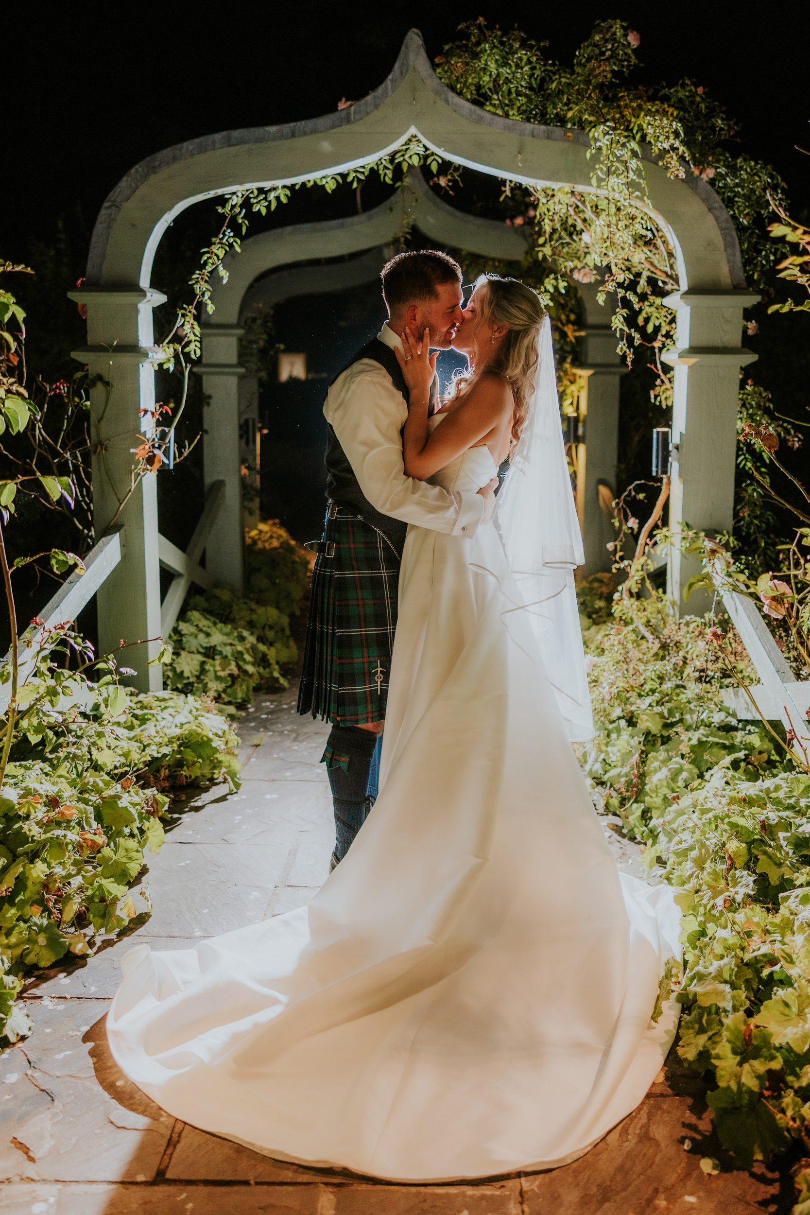 Bride in white gown and veil and groom in kilt kiss beneath a lit wooden archway at night, train trailing on the stone path.