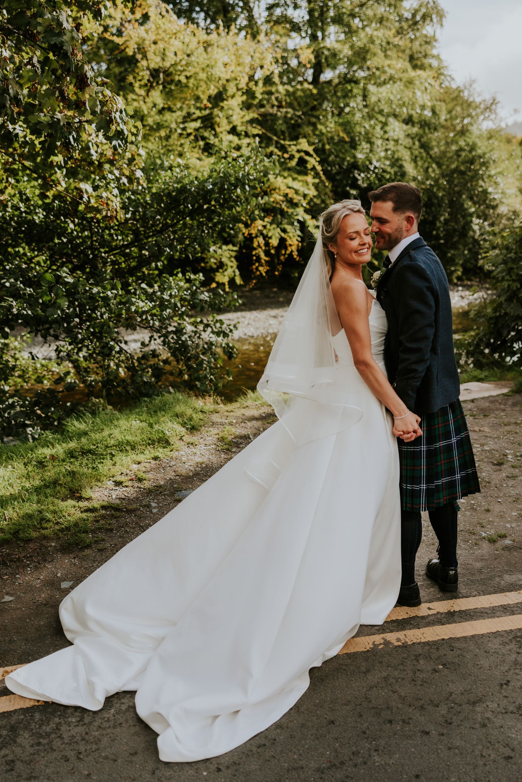 Bride in white gown and veil holding hands with groom in a kilt beside a riverside path and trees.