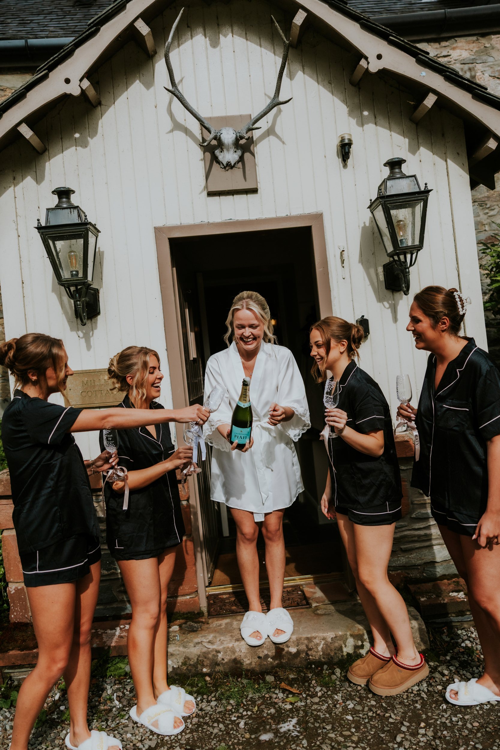 Bride in white robe pops a champagne bottle as bridesmaids in dark robes hold glasses and laugh outside a cottage door.