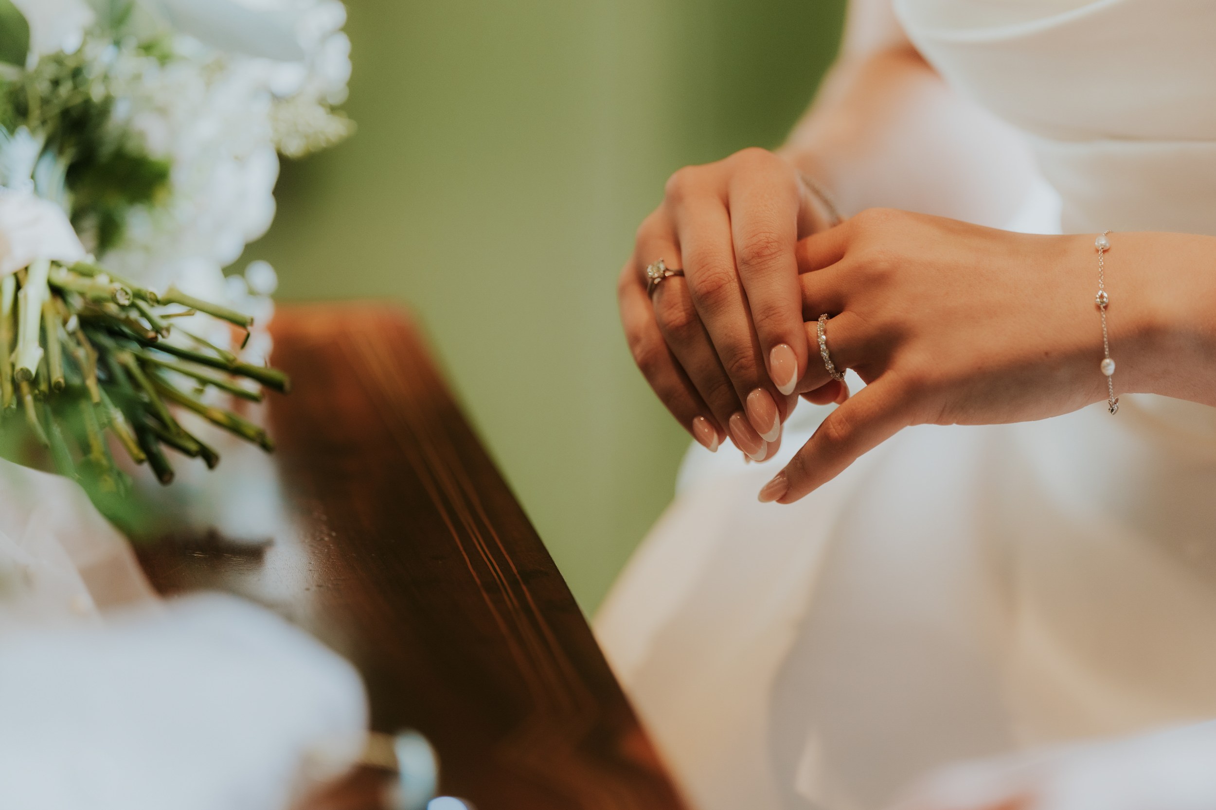 Bride's hands wearing engagement and wedding rings and a pearl bracelet, bouquet stems blurred at left.