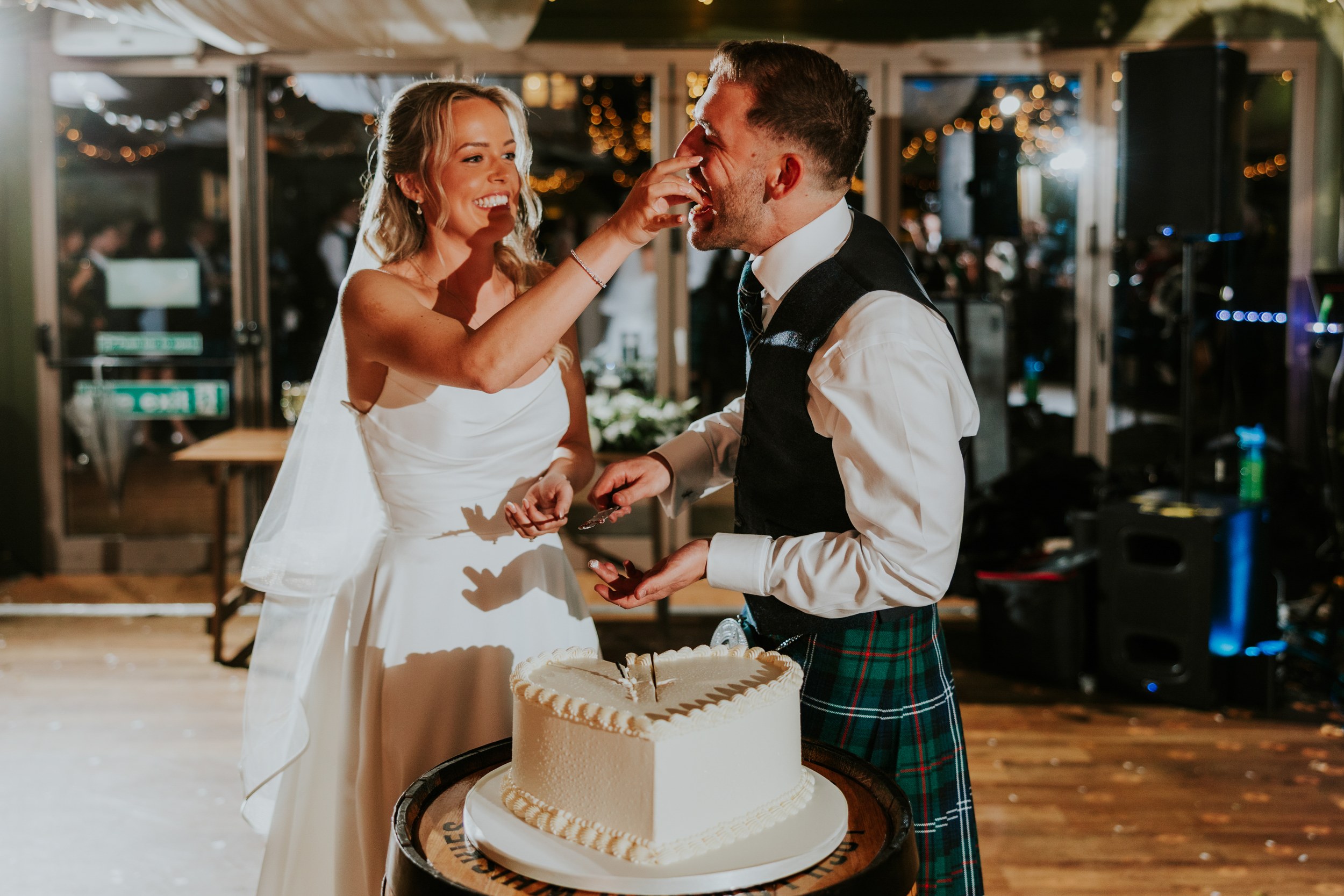 Bride smearing cake on the groom's face beside a heart-shaped wedding cake, guests and fairy lights behind them.