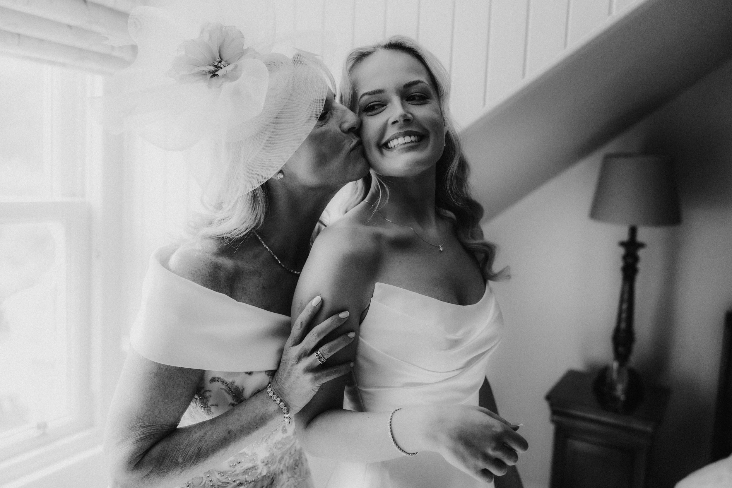 Bride smiling as an older woman kisses her cheek by a window during wedding preparations.
