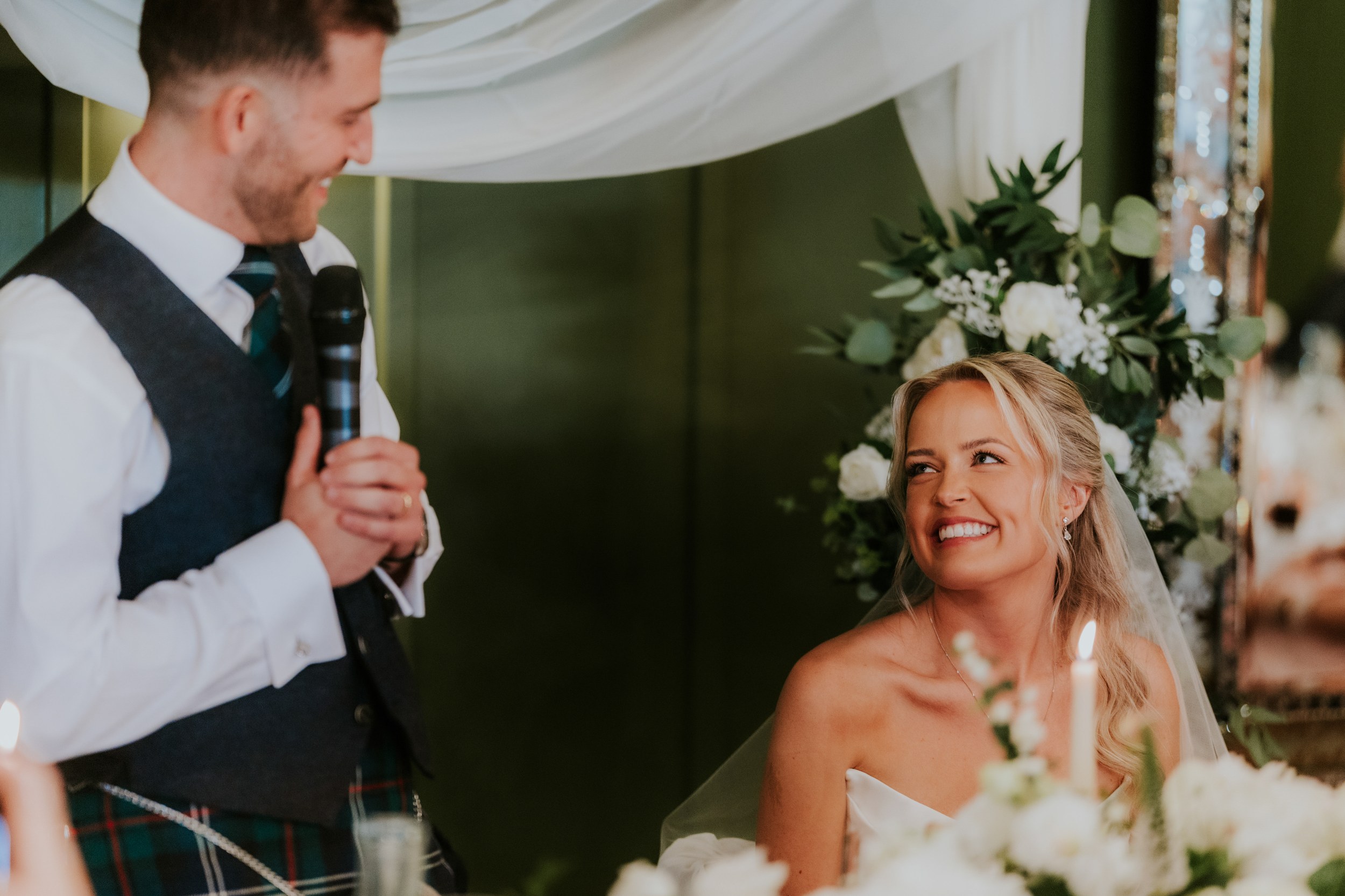 Bride smiling up at a man speaking into a microphone at the top table, with candles and floral arrangements nearby.