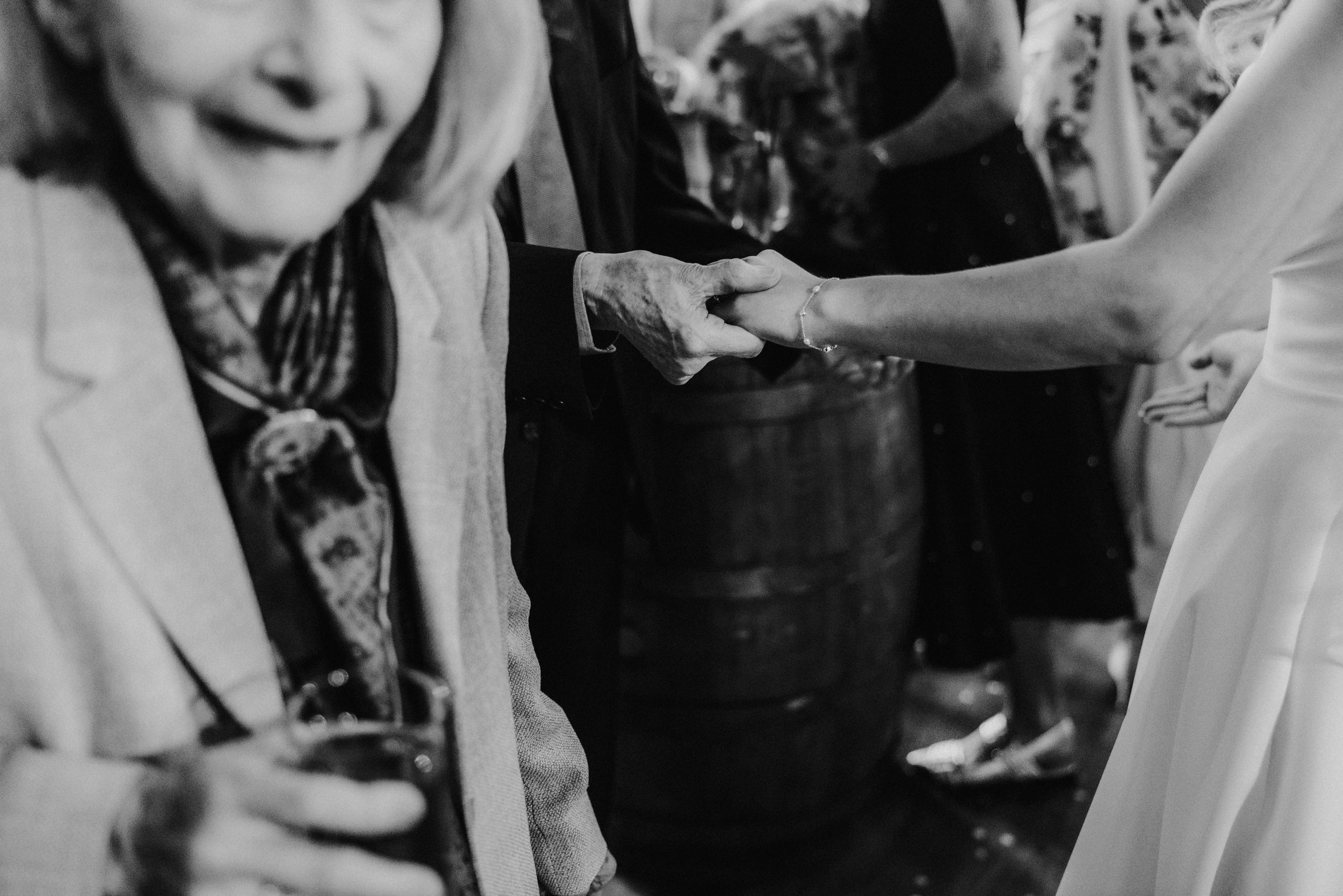 Close-up black-and-white of an elderly guest holding a woman's hand in a white dress during a drinks reception.