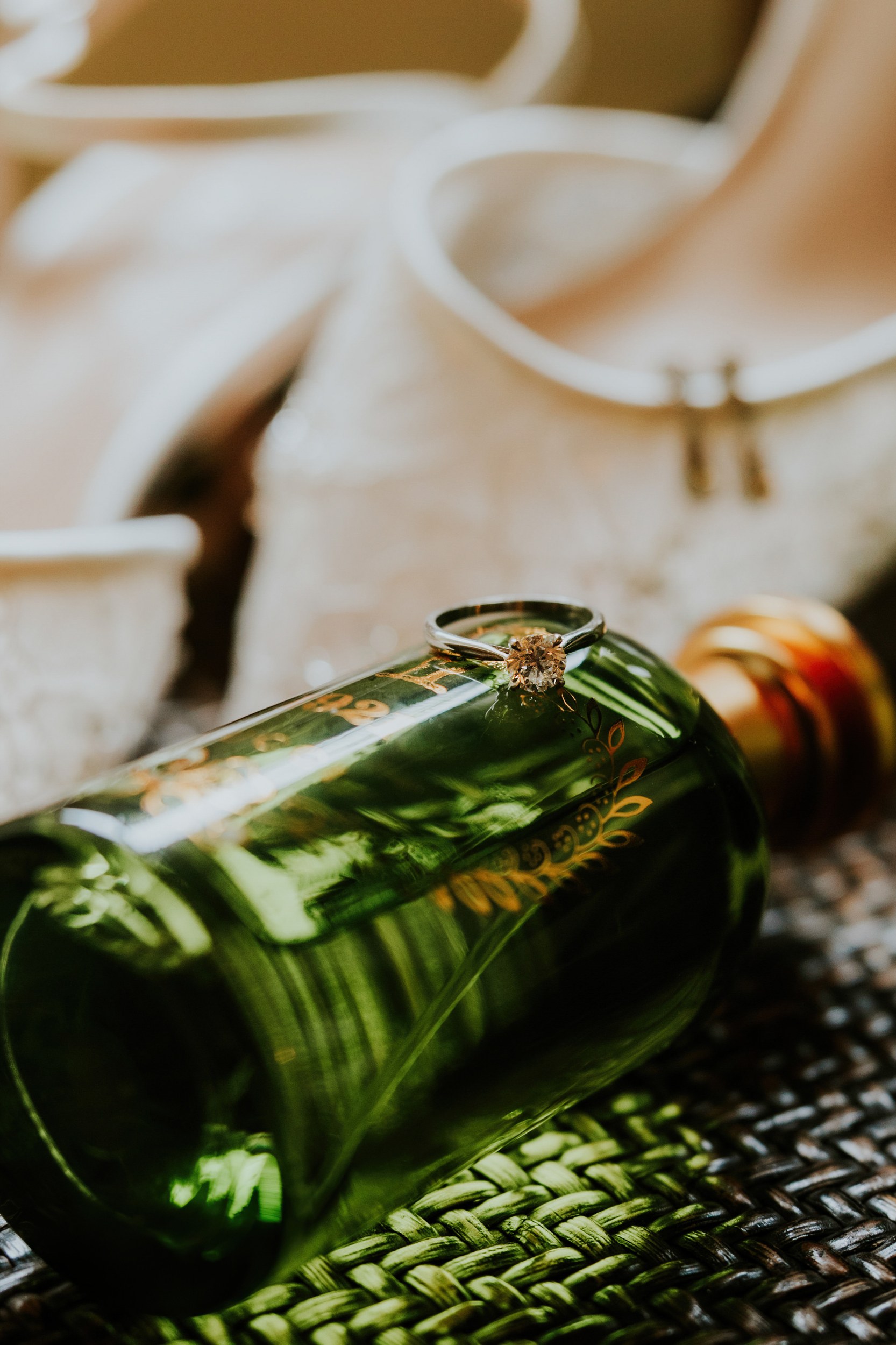 Diamond ring on a green decorative glass bottle with blurred bridal shoes behind.
