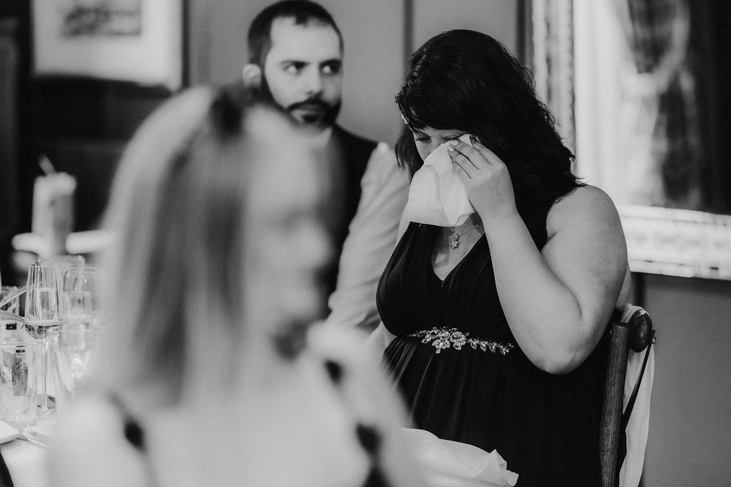 Female wedding guest wiping tears with a tissue at a table during speeches.